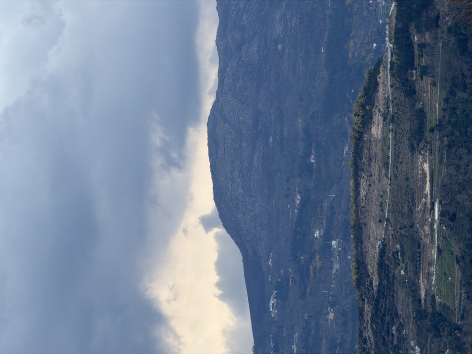 This image depicts a mountainous landscape within the city of Vila Real, Portugal. The upper portion of the frame is occupied by an overcast sky, characterized by dark grey stratocumulus clouds. A brighter, yellowish-white band of clouds is visible just above the horizon, suggesting indirect light.
A prominent, large mountain dominates the mid-background, its summit and upper slopes appearing dark blue-grey with visible rocky textures and sparse, scrubby vegetation. Below this, a series of successive hills descends towards the foreground. These hills are covered predominantly in dark green and brown vegetation, consistent with forested areas or dormant deciduous trees. Numerous white and light-colored buildings, identifiable as residential structures, are scattered across these hills, indicating a dispersed settlement pattern.
In the lower mid-ground, a winding asphalt road is visible, traversing a hillside with a gentle curve. Below this road and in the immediate foreground, terraced agricultural fields are discernible, displaying patches of green cultivation and exposed brown earth. No individuals are present in the scene. The environment is rugged, natural, and rural, with evident human intervention in the form of housing and agriculture.