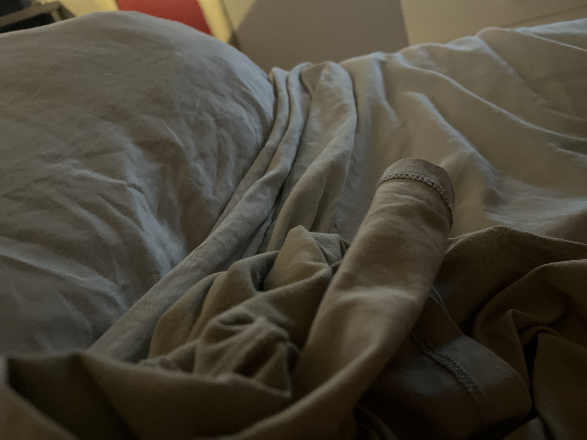 A close-up, low-angle shot shows a rumpled bed with light-colored sheets and a darker blanket. A grey t-shirt is partially visible in the foreground, draped over the blanket. The fabric of the t-shirt shows stitching along the hem. The lighting is dim, with soft shadows cast by the folds of the fabric. The scene appears to be indoors, in a domestic setting, such as a bedroom. There is no indication of location in Minsk, Belarus, or any specific city. The image focuses on textures and the play of light on fabric.