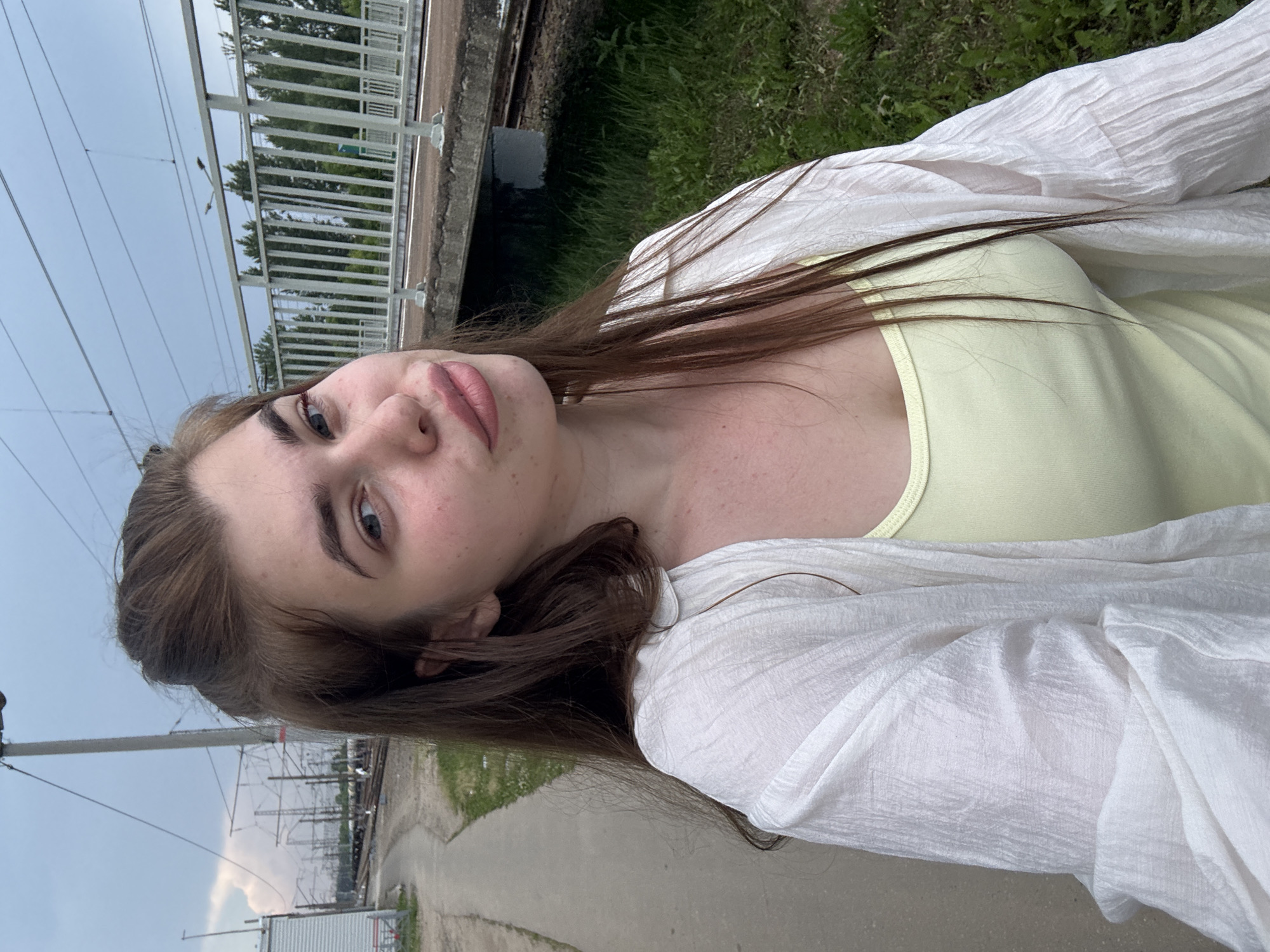 A young Caucasian woman with long, dark brown hair pulled back at the crown of her head is pictured from the chest up. She is wearing a light yellow tank top under a sheer white open-front shirt. The woman is standing on a concrete walkway next to a set of railroad tracks. A raised train platform with a white railing is visible to her right, with trees in the background. Overhead power lines and poles are present, suggesting an industrial or transportation area. The sky is overcast. The woman's face is turned towards the camera with a slight tilt of her head and a hint of a smile. She has light blue eyes and is wearing light makeup. Her skin appears fair with some freckles. The overall setting seems to be a train station or a railway yard in a city, potentially Rechytsa, Russia, given the context provided.