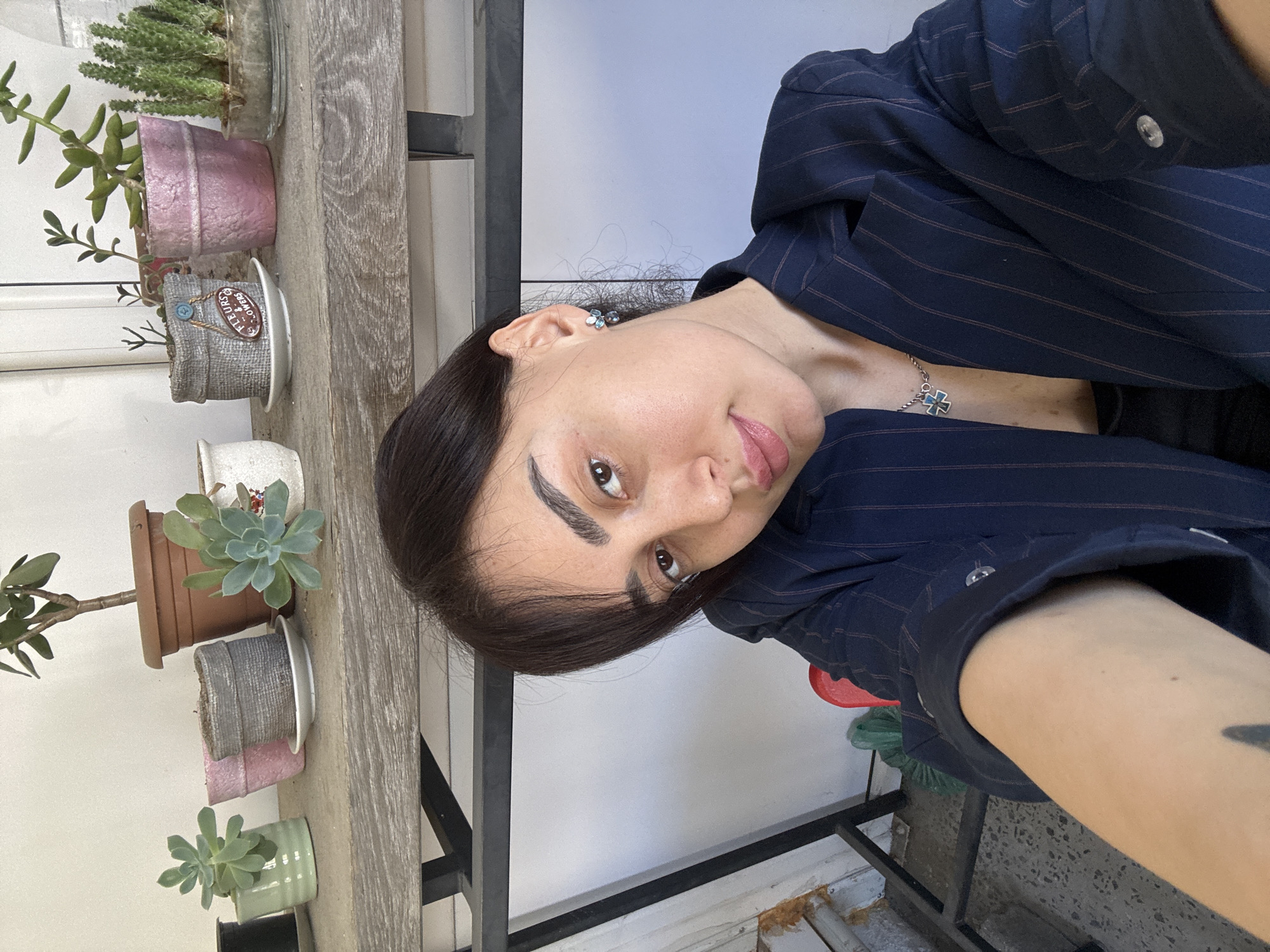 A selfie taken by a woman with dark hair, wearing a navy pinstripe blazer and a silver cross necklace. She is indoors, possibly on a balcony, judging by the row of potted succulents arranged on a wooden shelf behind her. The shelf is supported by a black metal frame. The background consists of a white wall. The location is likely in Tbilisi, Georgia, considering the style of architecture.