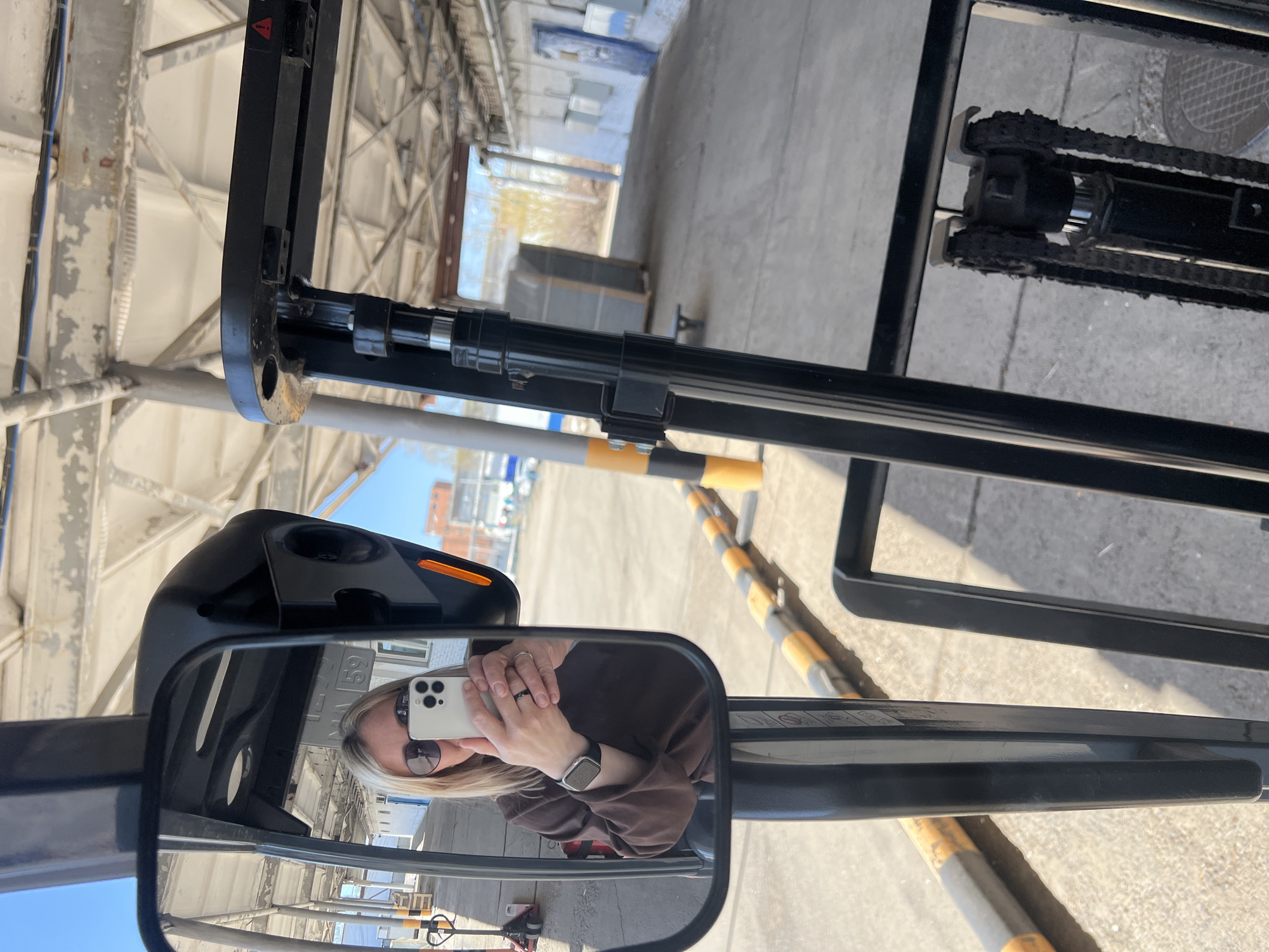 The image shows a reflection of a person in the side mirror of a forklift, within an industrial area of Chaikovsky, Russia. The person is fair-skinned with blonde hair, sunglasses, wearing a brown shirt, and holding a white smartphone to take a photo. Visible on their wrist is a watch.
The forklift is black and the mirror provides a view of the surrounding industrial environment, including an open warehouse or factory structure with a metal frame. Yellow and black striped bollards are situated near the forklift. In the background, one can see additional buildings, likely within the industrial complex. The sky is clear and sunny.