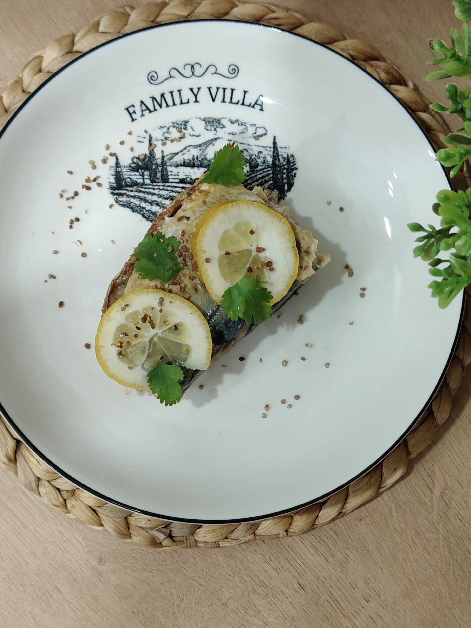 On a white plate with a black border and the words "FAMILY VILLA" written on it, rests a piece of fish on bread, garnished with lemon slices and cilantro. Small brown seeds are sprinkled around the food. The plate sits on a woven placemat on a wooden surface. A green plant is partially visible on the right side of the image.
