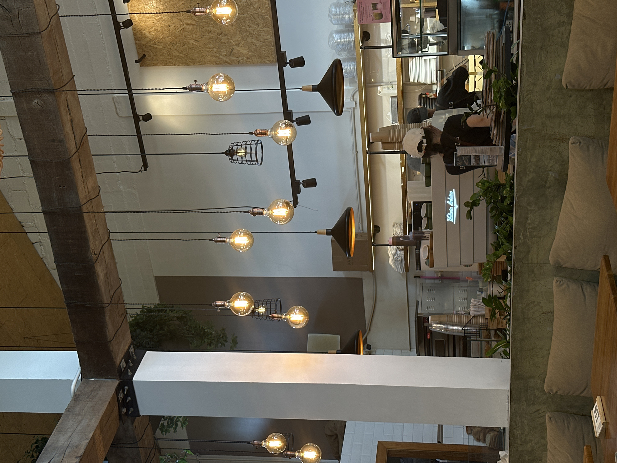 The image shows the interior of a cafe or restaurant in Choeng Thale, Thailand. The scene is characterized by rustic-industrial design elements, notably exposed wooden beams crisscrossed with electrical wires supporting decorative light bulbs and conical lamps. A serving counter is visible, attended by two individuals: one wearing a white cap and a black t-shirt, and another wearing a black hooded sweatshirt. The counter area contains equipment and supplies associated with a cafe, such as a coffee machine and disposable cups. Seating arrangements, including cushioned benches, are visible along one side of the frame. The decor includes potted plants.