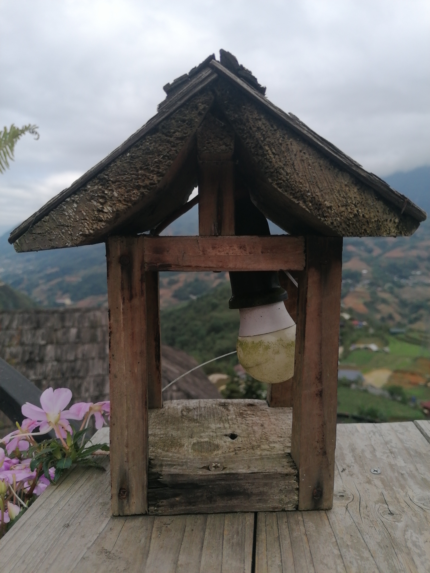 A wooden birdhouse-like structure sits on a weathered wooden surface, likely a deck or table, in Sa Pa Ward, Vietnam. The structure has a peaked roof made of textured wood, supported by four vertical posts connected by a horizontal beam. A lightbulb hangs inside the structure. To the left of the structure, a cluster of pink flowers adds a touch of color. In the background, a landscape of terraced fields and rolling hills stretches out under a cloudy sky. A tiled rooftop is partially visible in the background. The scene is rural, with the man-made structure contrasting with the natural landscape.