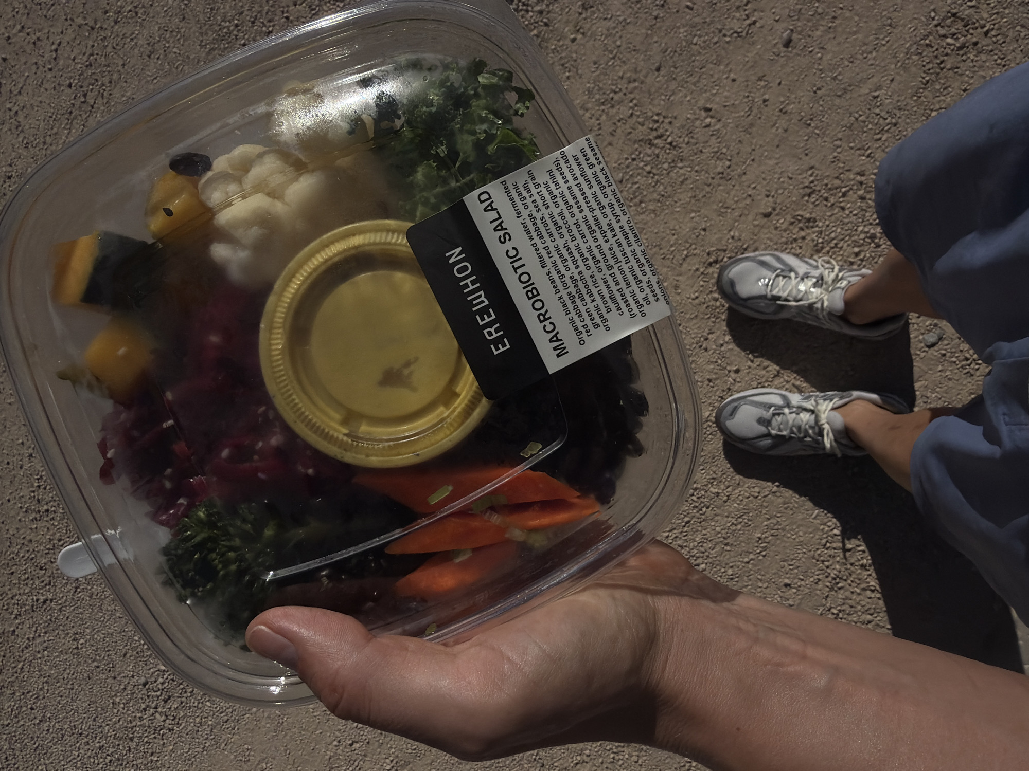 An overhead shot captures a hand holding a clear plastic container of a macrobiotic salad from Erewhon, likely on a gravel path. The salad contains black beans, red cabbage, carrots, kabocha squash, cauliflower, broccoli, and kale. A small yellow cup of dressing is inside the container. The person is wearing gray and white athletic shoes and blue shorts. The background is a gravelly surface, suggesting an outdoor, possibly arid environment. The image composition focuses on the salad and the person's hand and feet.