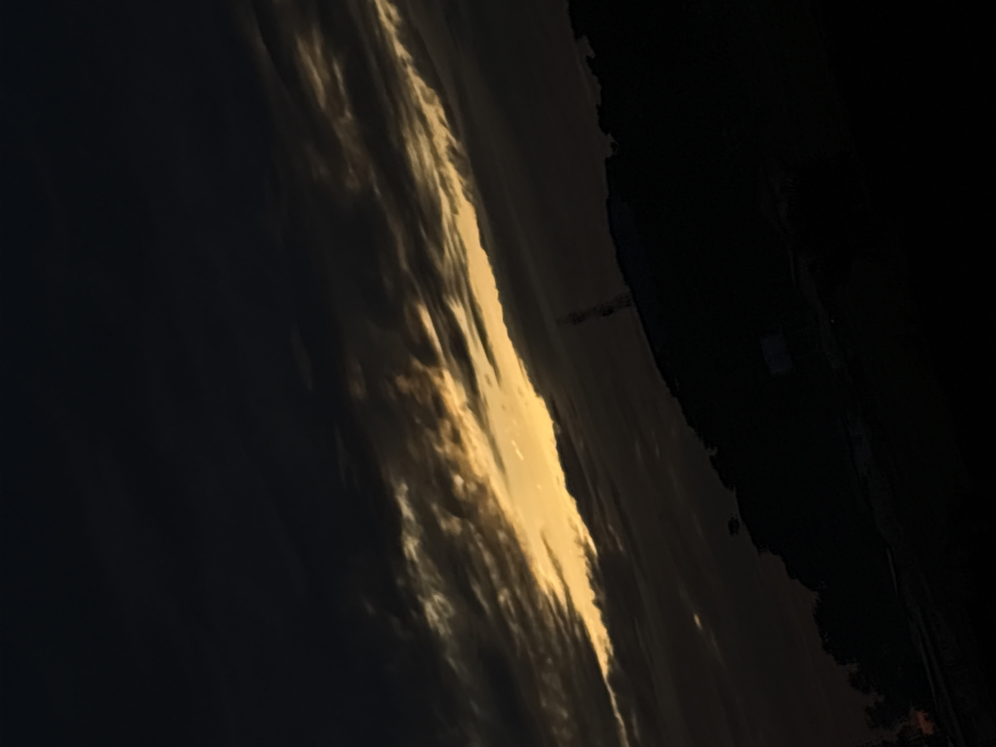 The image depicts a twilight sky, likely sunset, characterized by a dramatic interplay of dark clouds and a vibrant band of golden light. Heavy, dark grey clouds dominate the upper left and upper right portions of the frame, with their undersides and edges illuminated by a strong, warm light source. A diagonal swath of bright, golden-yellow light cuts across the mid-section of the sky, extending from the mid-left to the mid-right, indicating the position of the setting or rising sun behind the cloud cover.
Below the illuminated sky, the lower third of the image presents a dark silhouette of a landscape, presumably hills or a mountain range, consistent with the terrain around Bukuru, Nigeria. The silhouette reveals a jagged, irregular outline, suggesting dense vegetation or rugged topography. Prominently positioned on this silhouetted ridge, slightly to the left of the image's center, stands a tall, slender telecommunications tower, distinguished by its vertical structure and visible cross-members, indicating human infrastructure within the natural environment. The very bottom of the image is obscured in complete darkness, showing no discernible details of the immediate foreground. No people, discernible actions, or direct interactions are visible within the scene.