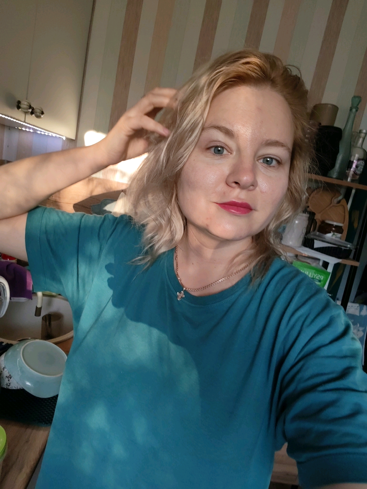 A blonde woman with blue eyes and bright pink lipstick is posing for a selfie. She is wearing a teal t-shirt and a delicate necklace with a small pendant. Her left hand is raised to her hair. She is standing in what appears to be a room with striped wallpaper in the background. There are shelves with various items, including bottles and jars, to her right. In the foreground to her left, a light blue bowl is visible. The lighting suggests it is daytime, with sunlight casting shadows on her shirt. The setting does not provide specific indicators to pinpoint the location within Izhevsk, Russia.