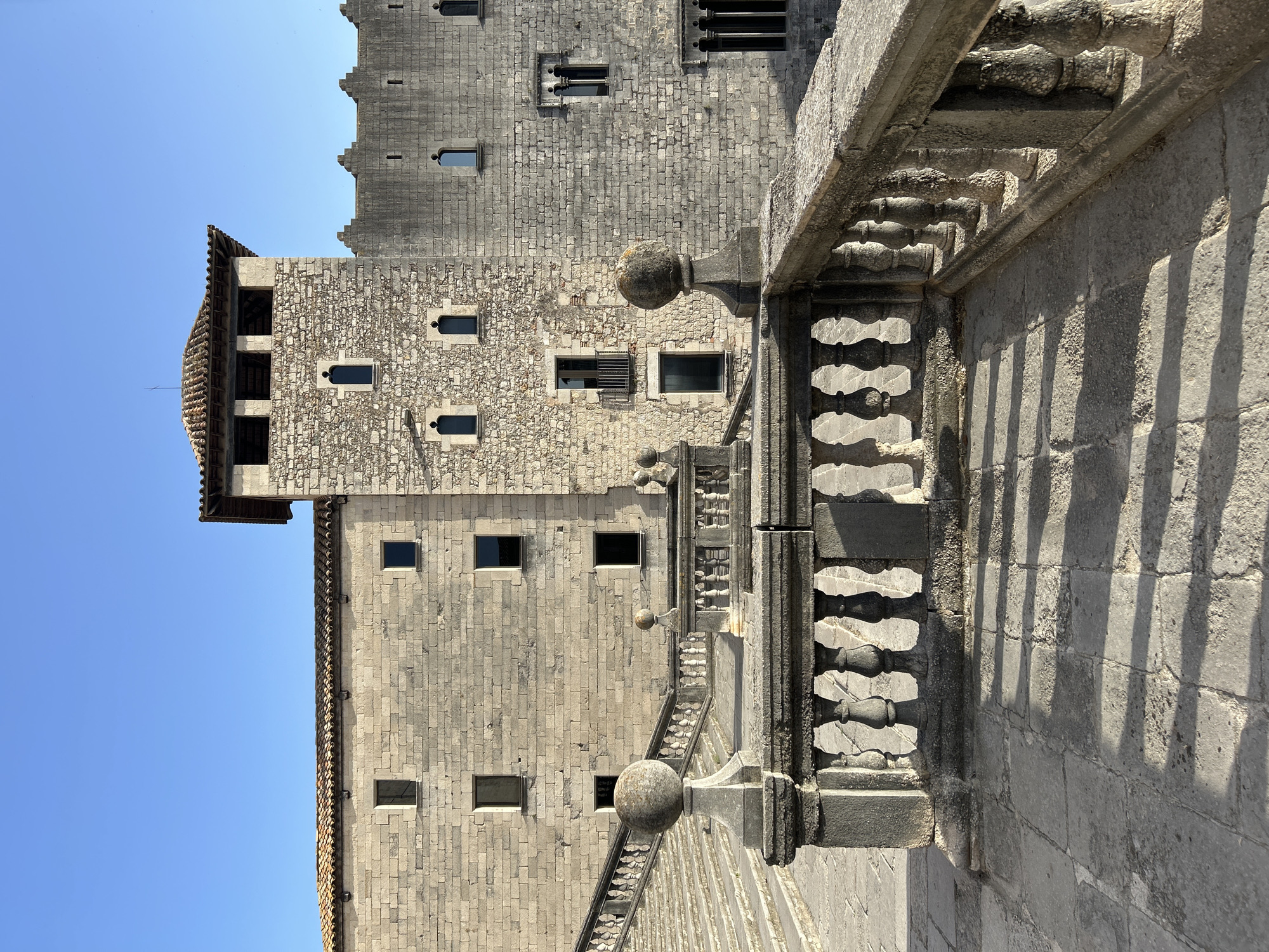 The image shows a stone building with a large stone staircase and a balustrade in the foreground, located in Girona, Spain. The building is constructed of large, rough-hewn stone blocks and features multiple windows. One section of the building has a prominent tower with a tiled roof. The staircase is wide and made of stone, with a stone balustrade featuring decorative balusters and finials. The ground in the foreground is also paved with large stone slabs, casting long shadows from the balustrade. The sky is clear and blue, indicating a sunny day. There are no people visible in the image. The scene appears to be a historical site or landmark.