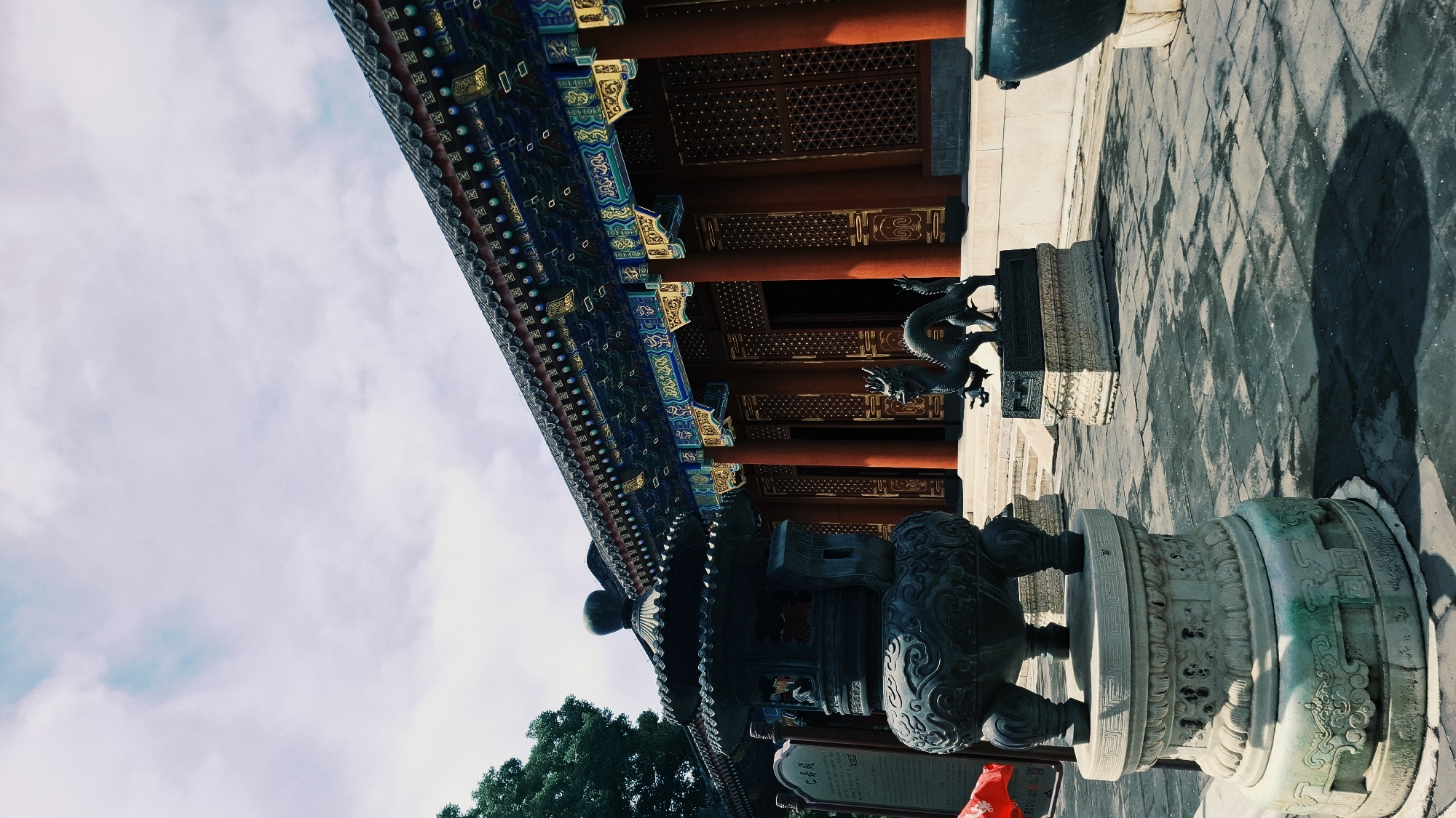 The image captures a detailed section of an ornate Chinese building in Haidian District, China. The building showcases intricate architectural details, including a multi-tiered roof with blue, green, and gold embellishments, supported by red pillars. A bronze dragon statue sits atop a stone pedestal. In the foreground, a large, decorative bronze incense burner on an intricately carved stone base is visible. The sky is partly cloudy. The ground is tiled with a grid-like pattern, marked by shadow.