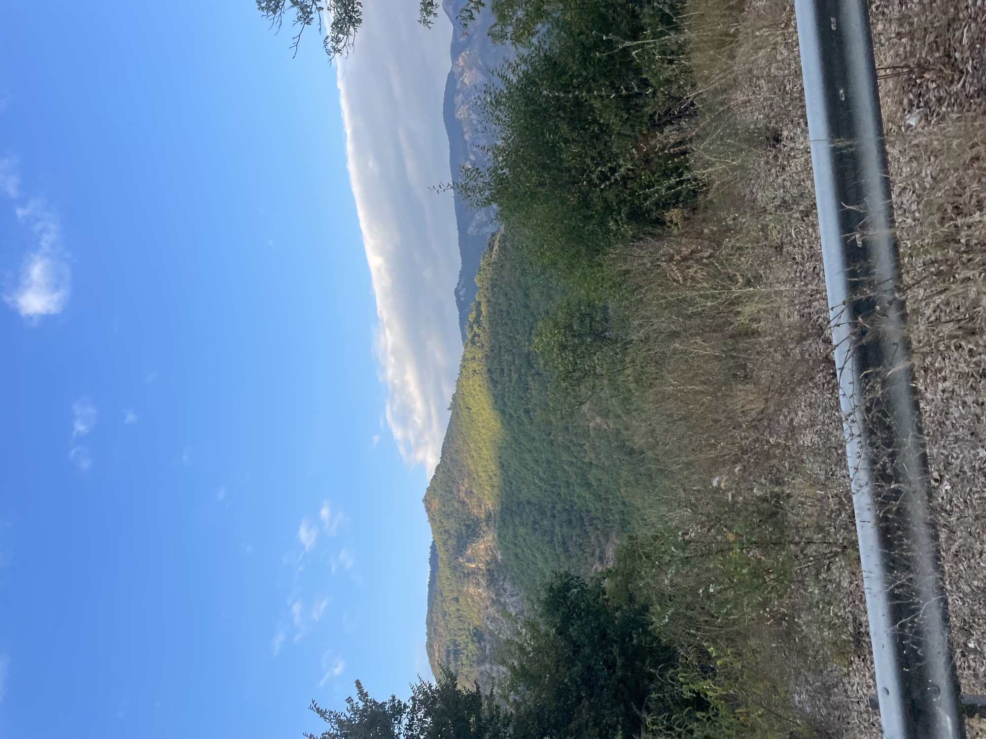 The image captures a scenic view of rolling, tree-covered mountains under a bright blue sky with scattered white clouds. The foreground features a grassy verge and a metal guardrail, suggesting a roadside perspective. The lush green of the trees contrasts with the rocky outcrops visible on the mountainside, and a distinct shadow divides the slope, indicating the position of the sun. The distant mountain ranges appear hazy and muted in color, fading into the background. The overall impression is one of a natural, undeveloped landscape. The location is identified as Yugovo, Bulgaria.