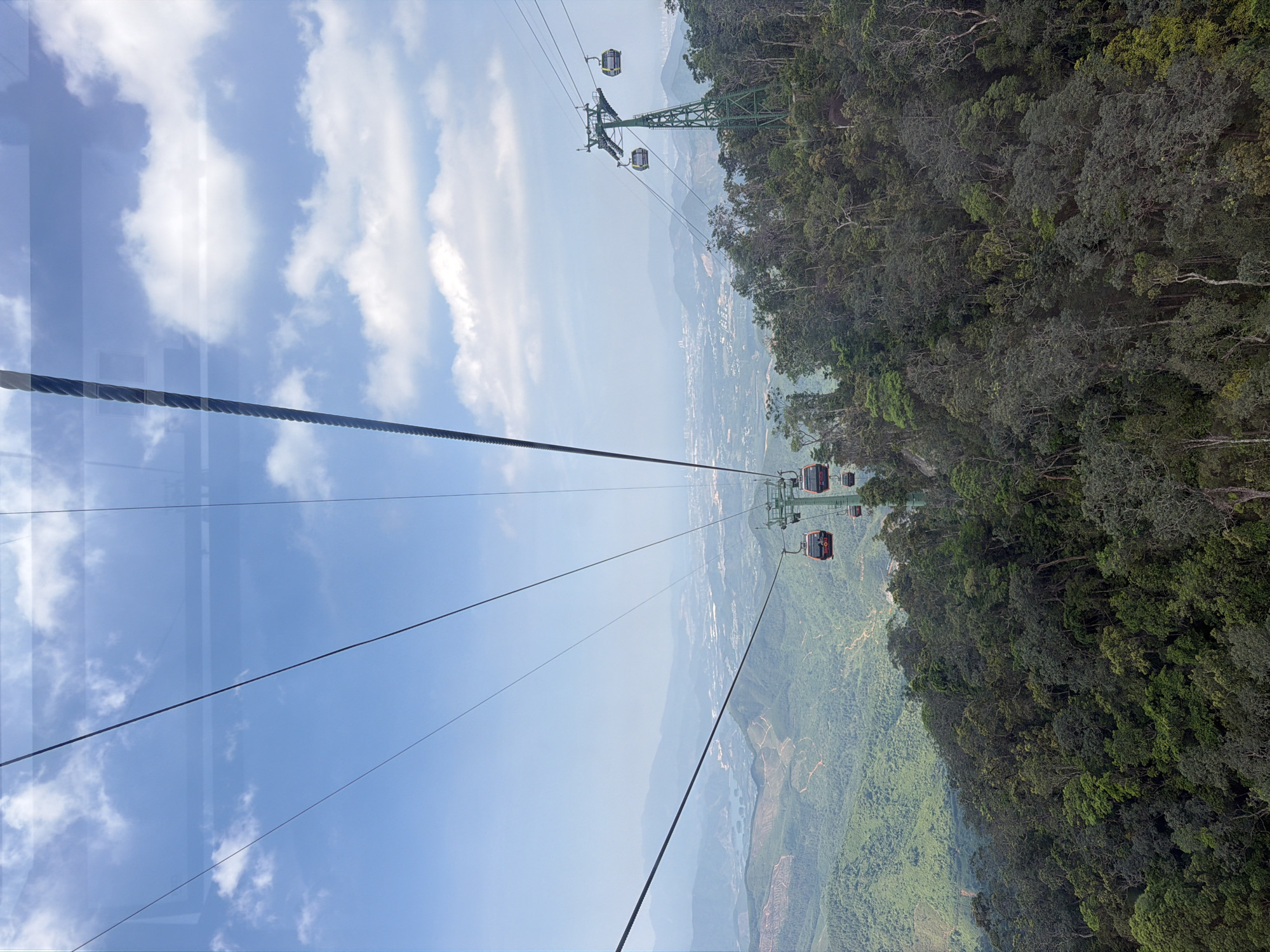 The image shows a cable car system in Đà Nẵng, Vietnam, likely Ba Na Hills. Several cable cars are visible, suspended from cables strung between green metal support towers. Below the cables is a steep hillside covered with dense forest. In the distance, there is a hazy view of the city and mountains. The sky is partly cloudy with a light blue hue. The photograph is taken through a window, with reflections visible.