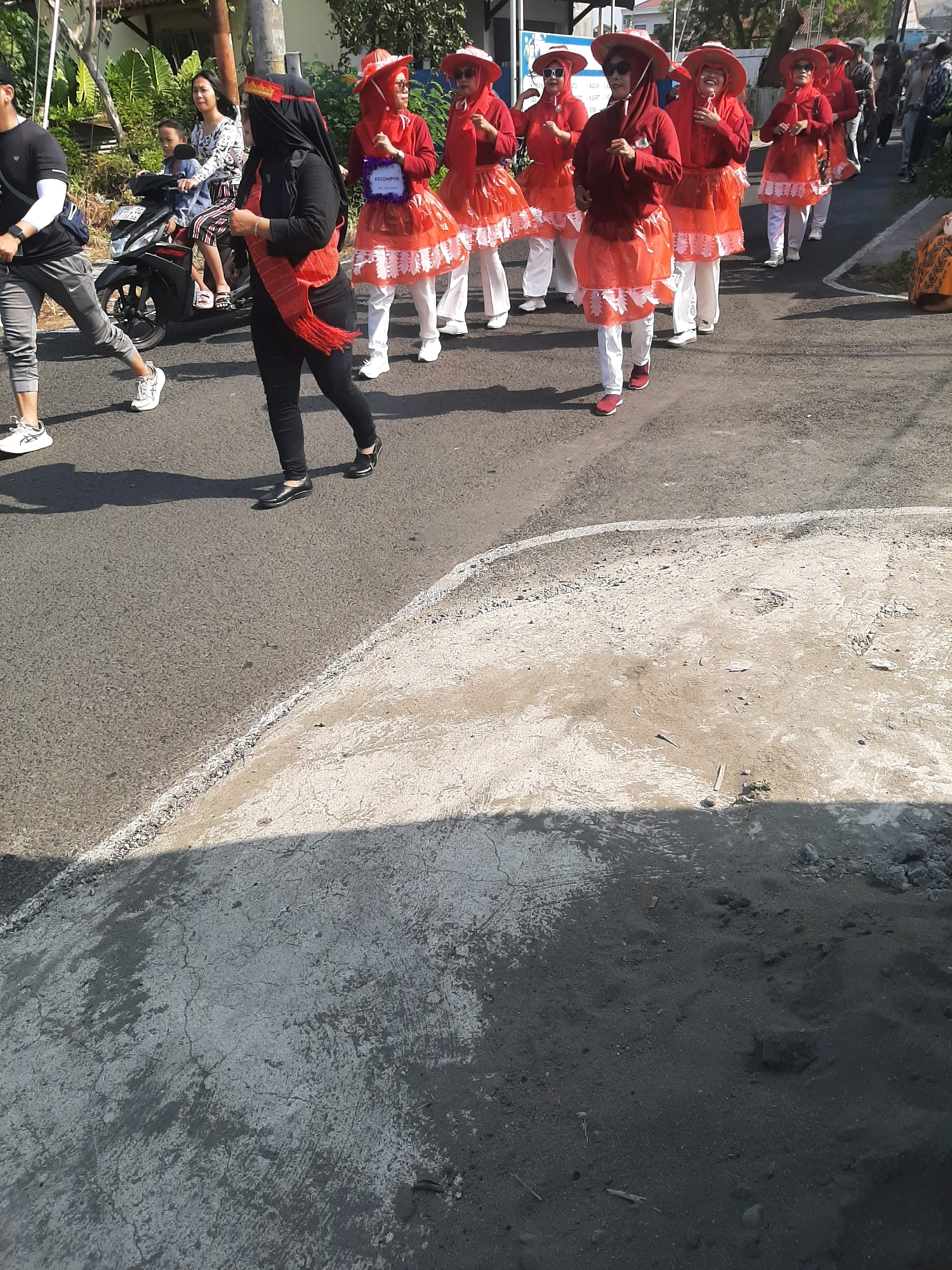A group of women in red and white costumes and hats are parading down a street in Sumampir, Indonesia. Some of the women are wearing hijabs. One woman in the foreground is wearing a black hijab and a red sash. In the background, a man is walking on the left side of the street, and a woman is riding on the back of a motorcycle with a child. The street is paved, and there is a sidewalk on the right side of the street with some people sitting down. The weather is sunny.