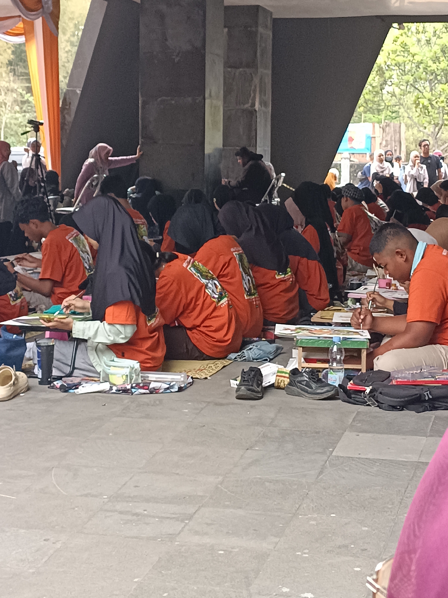 This image depicts an outdoor art event or competition held in Hargobinangun, Indonesia, likely in a public space with paved flooring and architectural elements. A large group of individuals, predominantly young adults and teenagers, are seated on the ground, engaged in drawing or painting. Many participants are wearing matching orange t-shirts with a design on the back, suggesting an organized event. Various art supplies are visible, including sketchpads, paints, brushes, water cups, and palettes. The environment features large, dark, concrete pillars and a grey, tiled floor. In the background, there are hints of tents or canopies in orange and white fabric, and foliage from trees. The overall atmosphere appears to be one of focused activity and collective participation in an artistic endeavor.