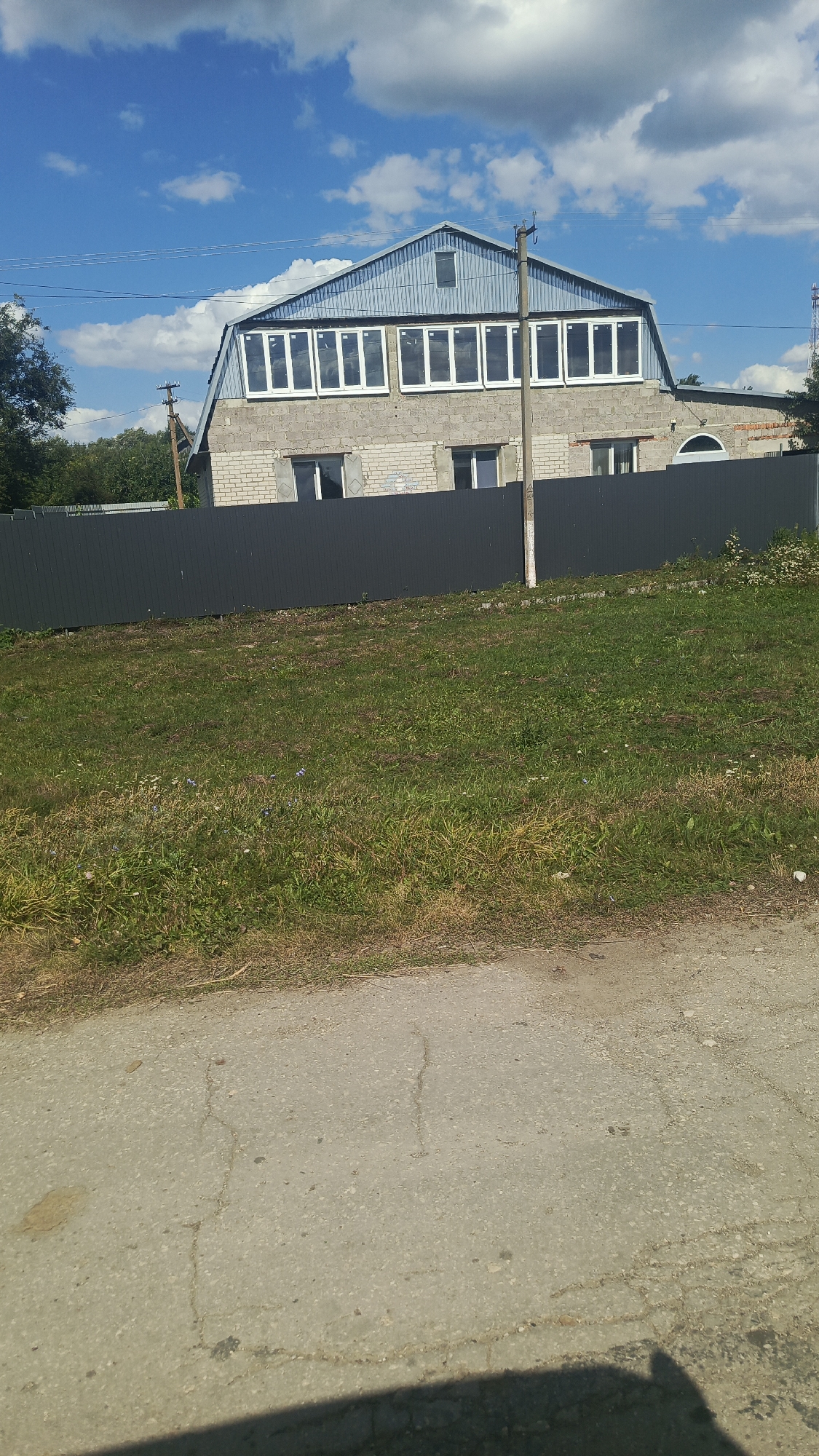 The image shows a building with a steeply pitched, light blue metal roof and a second story with large, rectangular windows. The first story of the building is constructed of light-colored bricks and has several smaller windows and a partially visible arched window. A dark gray metal fence runs across the front of the property, obscuring the lower portion of the building and yard. The area in front of the fence is covered in green grass with some patches of dry grass and small wildflowers. A paved surface, likely a road or path, is visible in the foreground. In the background, a utility pole with wires is situated near the building. The sky is blue with scattered white clouds. The overall scene appears to be a residential or commercial property in a semi-rural or suburban setting. The location is identified as Октябрьский Городок, Russia. There are no people visible in the image.