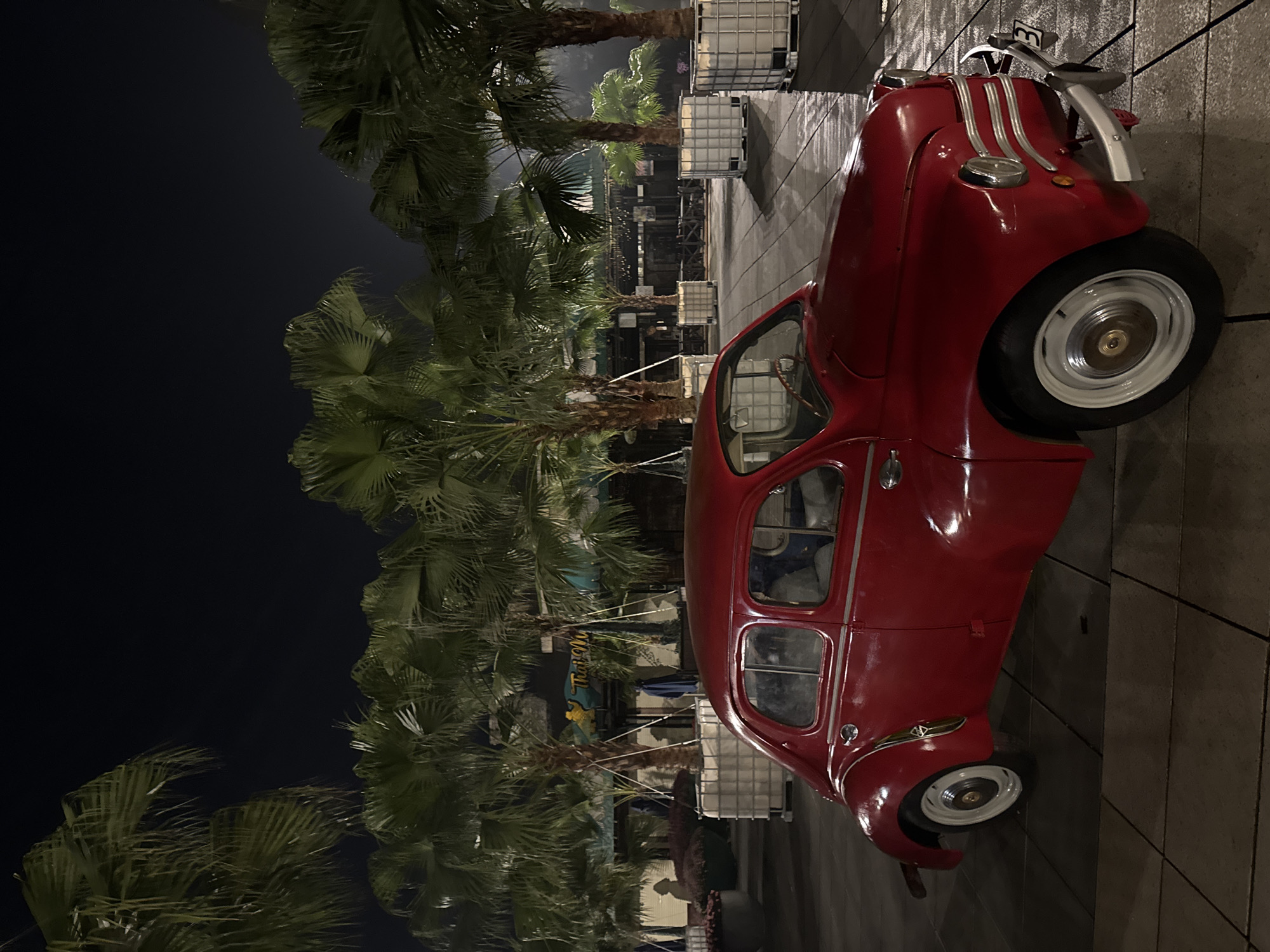 The image, taken at night in Đà Nẵng, Vietnam, features a striking red, vintage-style car as its primary subject. The vehicle, which exhibits classic design elements like a rounded body, prominent chrome grille, round headlights, and white-wall tires or white hubcaps, is parked on a tiled, paved surface.
The setting is an outdoor area characterized by numerous tall palm trees that line the background, their fronds visible against the dark night sky. Artificial lighting illuminates the scene, highlighting the green foliage of the trees and the glossy finish of the car. In the mid-ground, behind the car and among the palm trees, several large, white, cubic storage containers, likely IBC totes, are visible, suggesting an event space, temporary installation, or utility area. Further back, indistinct structures or buildings can be discerned.
No people are visible in the photograph, and the car appears to be a stationary display or simply parked. The time of day is clearly night, with no indications of adverse weather. The combination of the classic car and tropical palm trees creates a unique, atmospheric, and perhaps nostalgic feel. A partial sign bearing the word "Thai" is faintly visible between some palm trees in the background.