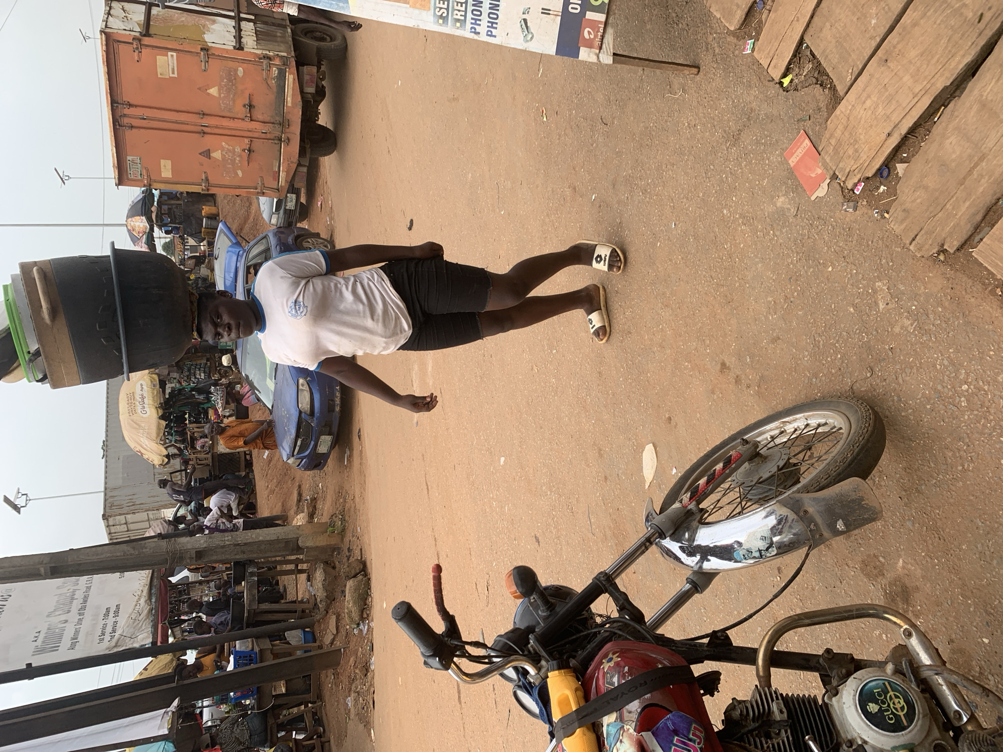 A young Black woman walks down a dirt road in Shagamu, Nigeria, carrying a stack of large dark pots balanced on her head. She is wearing a white t-shirt with a blue trim around the collar and sleeves, and black shorts. Her feet are clad in white and tan flip-flops. She is looking towards the camera. In the foreground, a red motorcycle is parked on the left side of the frame, with its front wheel turned to the right. The motorcycle has a chrome fender and handlebars, and a black engine. The woman is walking away from a marketplace, with stalls and people visible in the background. A large orange truck is parked on the right side of the road, and a blue car is visible between the woman and the truck. The sky is overcast.