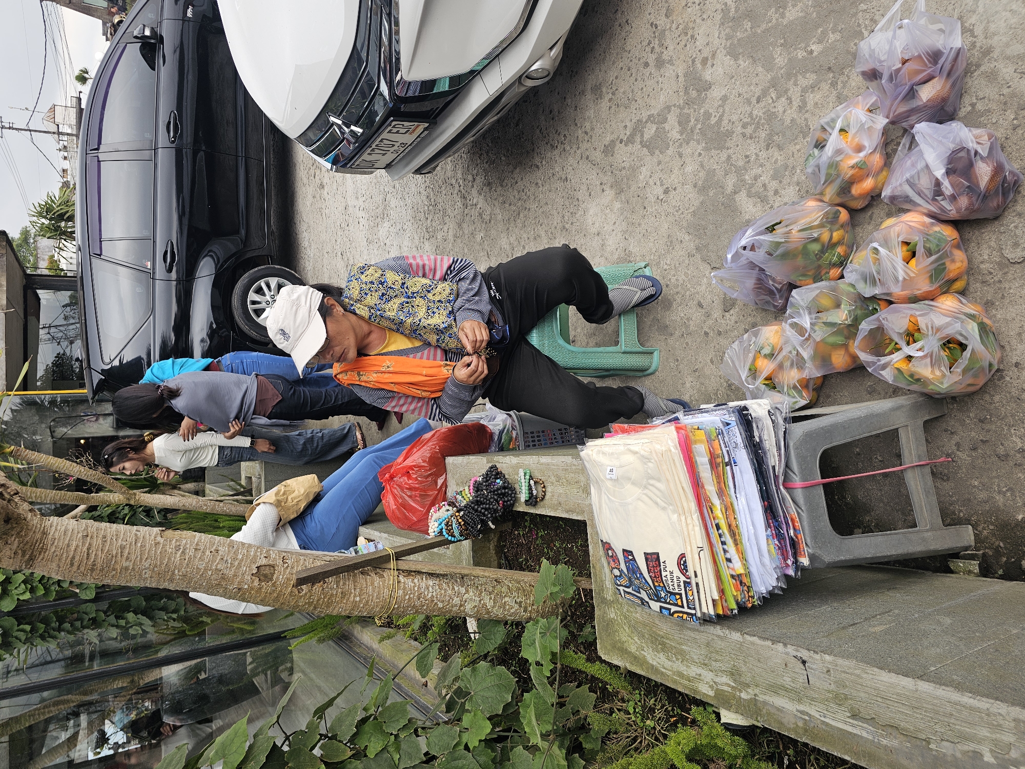In Kintamani, Indonesia, a woman sits on a green plastic stool beside a pile of t-shirts stacked on a gray plastic stool. She wears a white cap, an orange scarf, and a striped shirt with black pants. Next to her are several plastic bags filled with oranges. In the background, a black SUV and a white car are parked on a paved surface. To the left, lush green plants grow along a concrete wall. Three other women stand near the t-shirt pile, one wearing a white long-sleeved shirt and blue pants, another in a gray shawl and dark pants, and a third in a blue top.