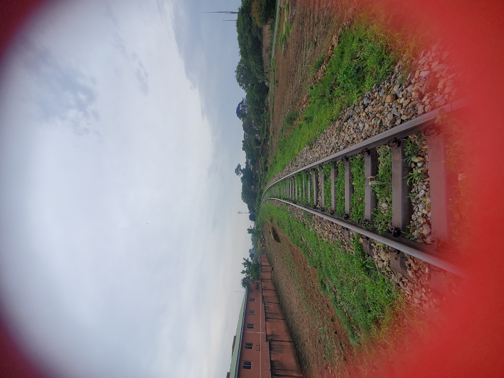 An image of a railway track curving to the left, receding into the distance under an overcast sky. The track is made of metal rails on wooden sleepers, surrounded by gravel and grass. To the left of the track is a low, reddish-brown wall topped with barbed wire, behind which is a long, multi-windowed building with a green roof. Further in the distance on the left, trees and some buildings are visible. To the right of the track, there are fields with visible rows of crops, and more trees and some structures in the background. The foreground is obscured by a large, blurred red oval shape. There are no people or animals visible in the image. The scene depicts an outdoor environment in a semi-urban or peri-urban setting. The image is likely taken in Jos, Nigeria, due to the building architecture, vegetation, and general landscape.