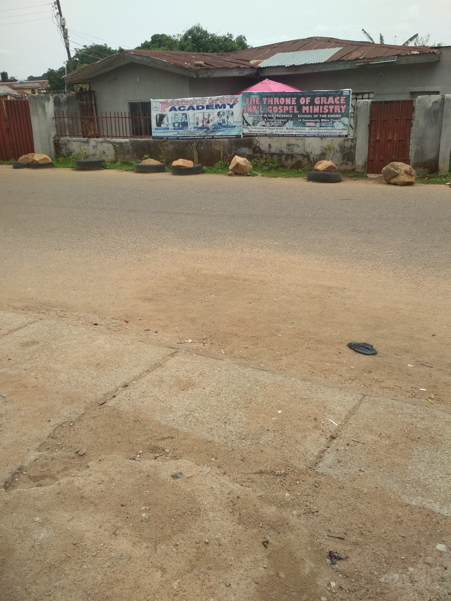 The image captures a street scene in an urban area of Unknown, Unknown. A one-story building is visible, with two signs attached to the fence: one for "Precious Stone Academy" and another for "The Throne of Grace Int'l Gospel Ministry." The fence is composed of concrete pillars and red metal bars. Large rocks rest on top of old tires in front of the fence. The road and adjacent sidewalk are worn and show signs of disrepair. A single black sandal lies on the ground near the sidewalk. The sky is overcast.