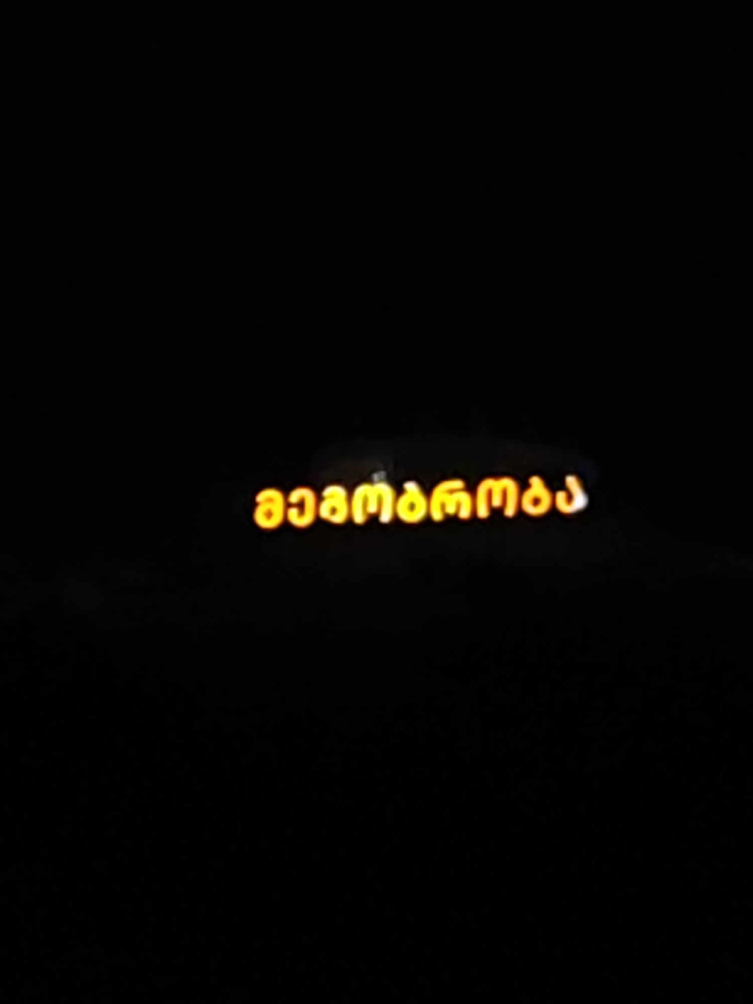 The image shows the word "მელომანია" (Melomania) written in bright orange Georgian script against a black background. The script is illuminated and appears to be neon or backlit. The text is centered horizontally and located slightly above the vertical center of the image. There are no people, specific objects, or environmental details visible in the image. The context of the word "Melomania" suggests a connection to music or a passion for music. The image does not provide any information about the city of Tbilisi, Georgia, or its specific location within the city.