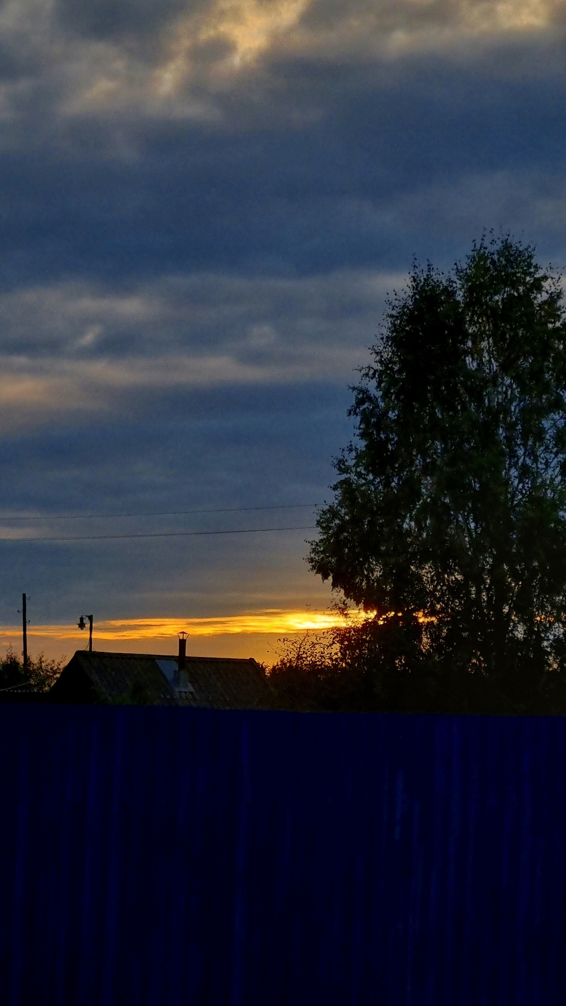 A vertical, close-up shot captures the dramatic sky and silhouetted landscape of Vysokoe, Russia, during twilight. The sky is a dramatic blend of deep blues and grays, with streaks of warm orange and yellow piercing through from the setting sun. A large, dark tree dominates the right side of the frame, its branches a stark silhouette against the vibrant sky. In the lower left quadrant, the dark silhouette of a house with a chimney is visible, nestled amongst other dark trees and foliage. A utility pole with what appears to be a light fixture stands near the house. The bottom portion of the image is obscured by a dark blue, textured area, possibly a fence or a draped cloth. The overall impression is one of a serene, rural evening.