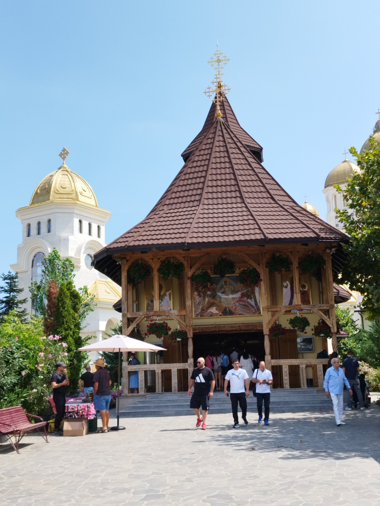 The image depicts a sunny outdoor scene in front of two Orthodox churches in Bucharest, Romania. A large, brown-roofed church with wooden architectural elements and painted murals is in the foreground. In the background, a white church with a prominent golden dome and a tall bell tower stands out. Several people are walking in front of the main church entrance, and others are seen near a market stall with an umbrella. The ground is paved with stone, and lush green trees and pink flowers surround the buildings. The sky is clear and blue.