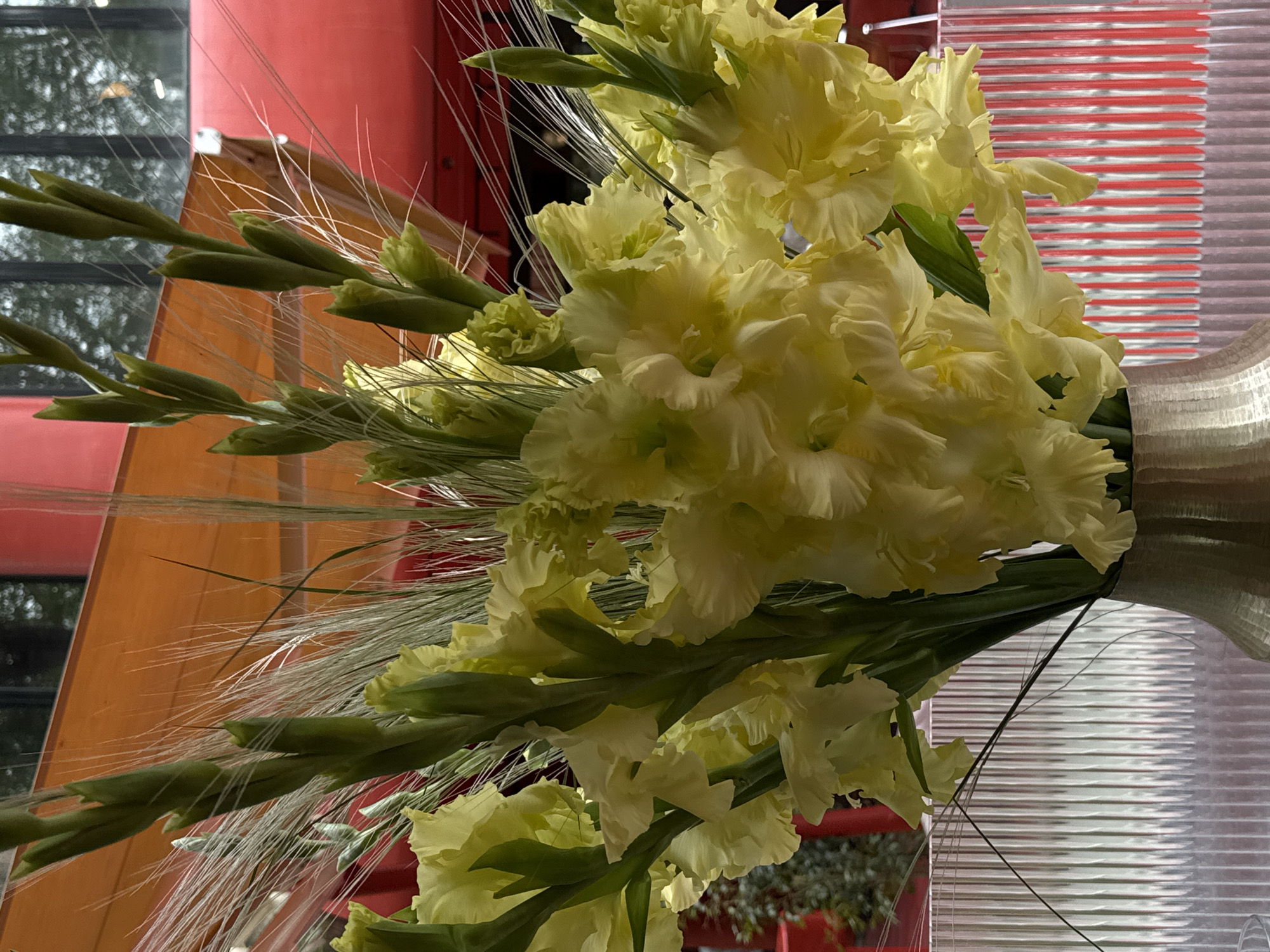 A bouquet of pale yellow gladiolus flowers, interspersed with green buds and wispy wheat-like strands, is displayed in a silver-colored vase. The background shows a partial view of a red architectural structure through a window or partition. This photo was taken indoors in the city of Yerevan, Armenia.
