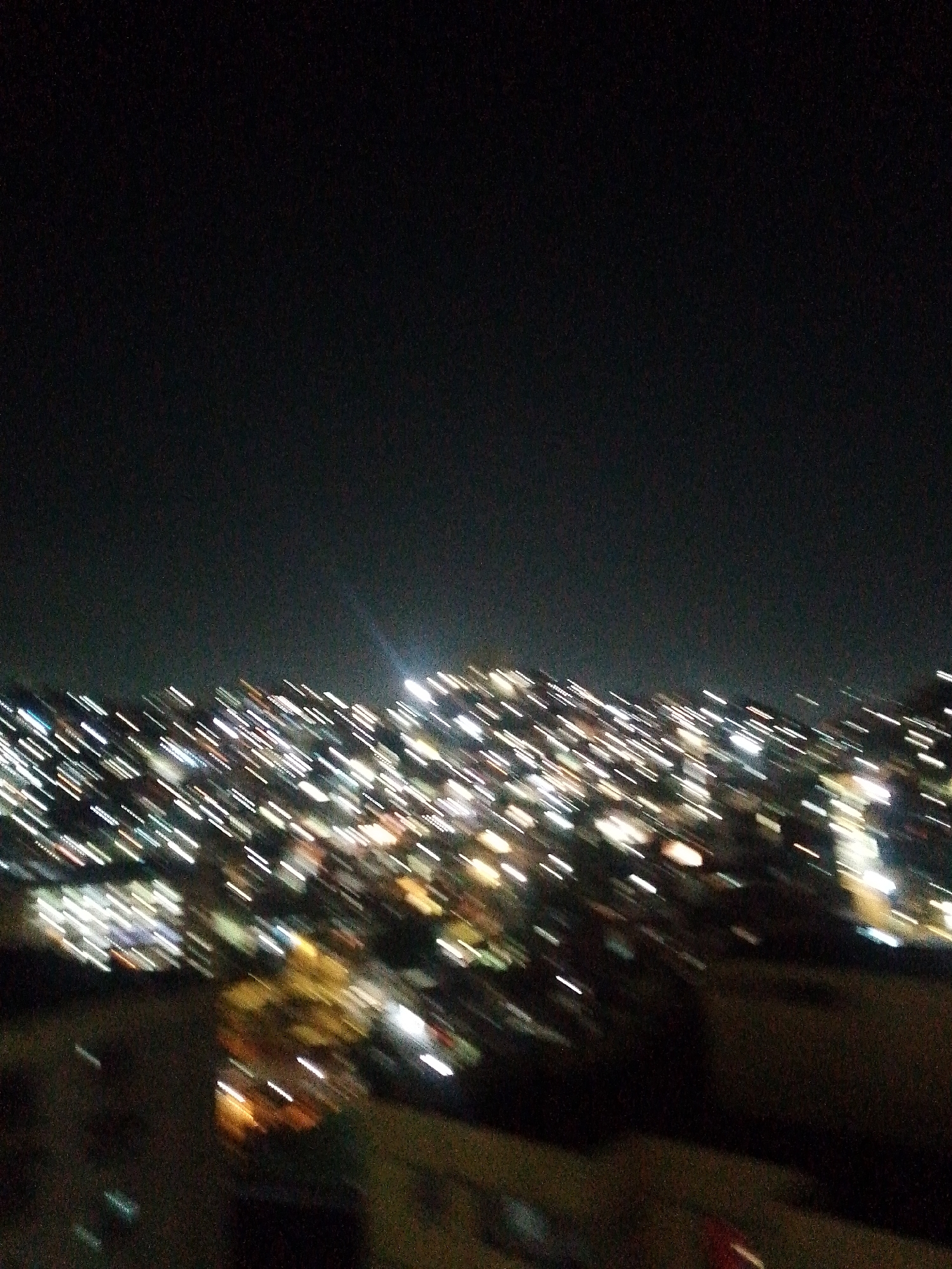 The image depicts a night view of Caracas, Venezuela, characterized by numerous blurred lights scattered across the city, likely due to camera motion or long exposure. The sky is dark, contrasting with the concentration of lights below, suggesting a dense urban landscape. There is no evidence of any specific people or actions visible in the image. The location is generalized based on the context provided.