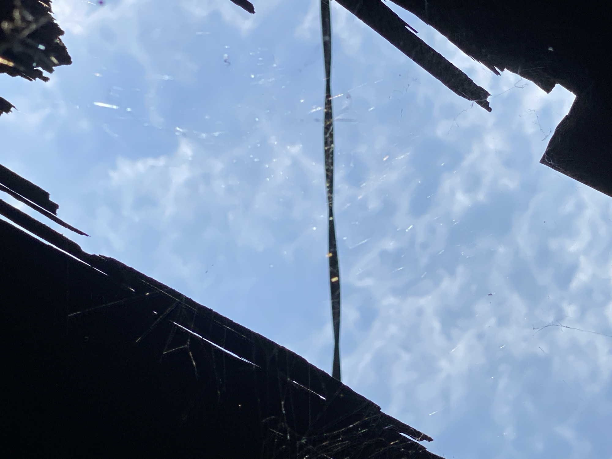 The image shows a close-up, upward-looking view through a broken roof or structure, revealing a bright blue sky with scattered white clouds. Dark, jagged silhouettes of the broken roof edges frame the sky, with thin strands of what appears to be spiderwebs visible across the opening. A horizontal, dark line, possibly a wire or debris, is stretched across the middle of the frame. The overall impression is one of decay and exposure to the elements. The location in Banjarmasin, Indonesia, suggests a tropical climate, which might explain the presence of spiderwebs and the bright sky. The scene depicts a damaged structure, possibly a dwelling or shed, with its roof partially collapsed, allowing a view of the sky. No people or specific actions are depicted.
