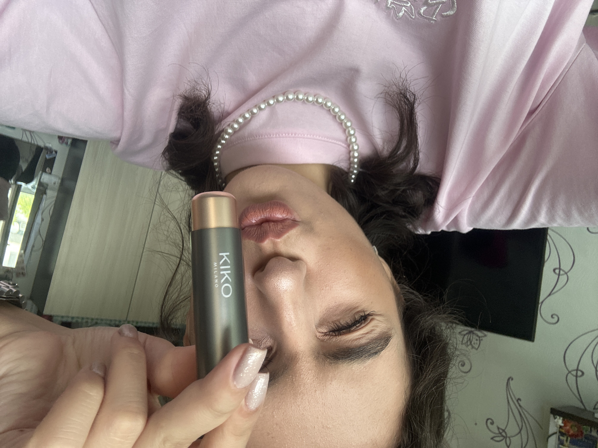A photograph depicts a woman from an upside-down perspective. She is holding a Kiko Milano product in her hand. The woman is wearing a pink shirt with a pearl necklace. The location is within a building, presumably in Unknown, Unknown. A wall with floral wallpaper can be seen in the background.