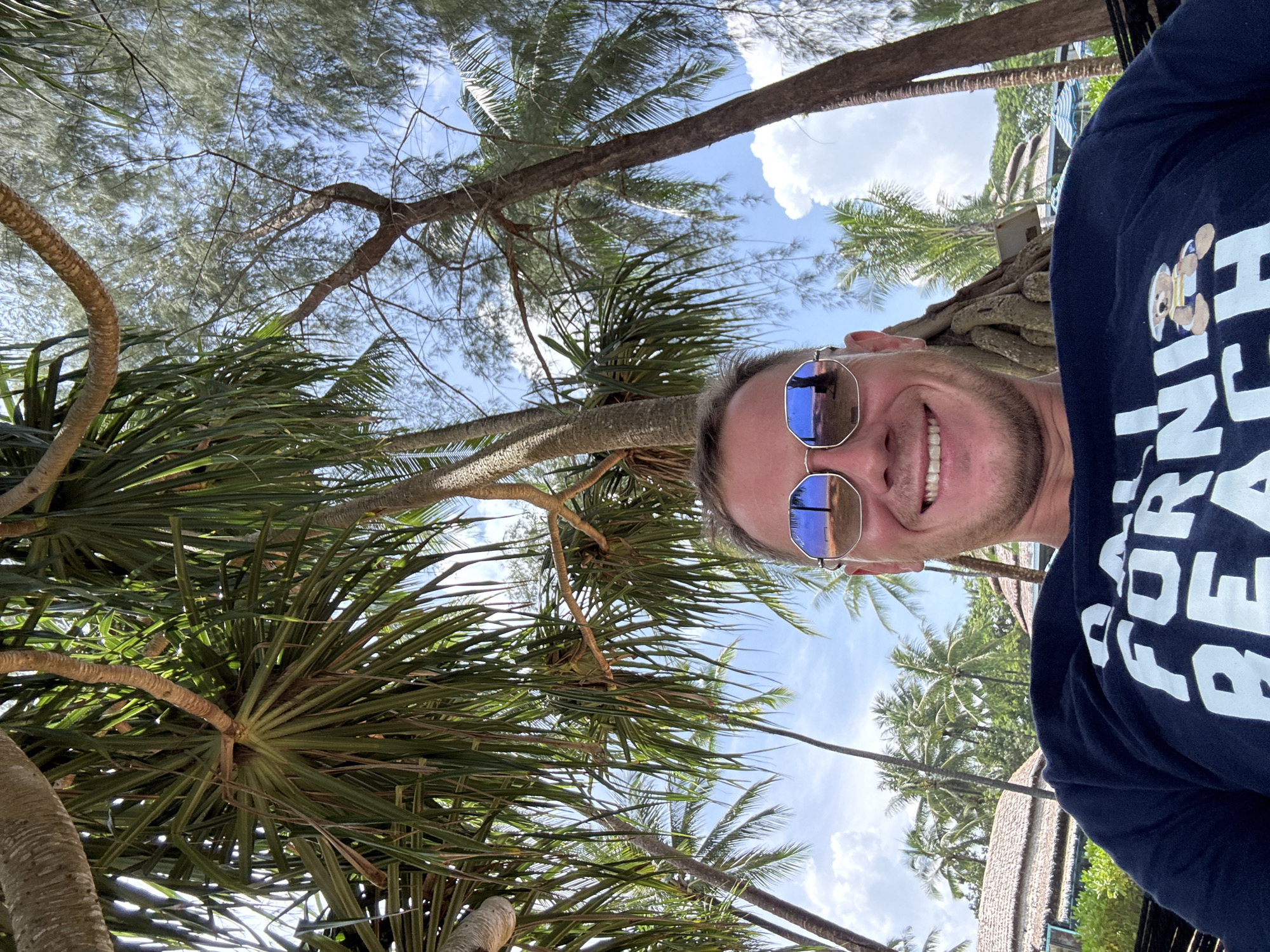 In Ban Khuk Khak, Thailand, a man in his 30s is lying in a hammock, looking at the camera. He is wearing mirrored sunglasses and a dark blue t-shirt with white lettering that appears to say "FORNI" and "BEACH". A cartoon bear is depicted on his shirt. The man is smiling and his face is framed by palm trees and other foliage. In the background, there are thatched-roof buildings, umbrellas, and more palm trees, indicating a resort or beach setting. The sky is blue with scattered white clouds. The reflection in the man's sunglasses shows a palm tree and a bench on a sandy area, further confirming the tropical beach location.