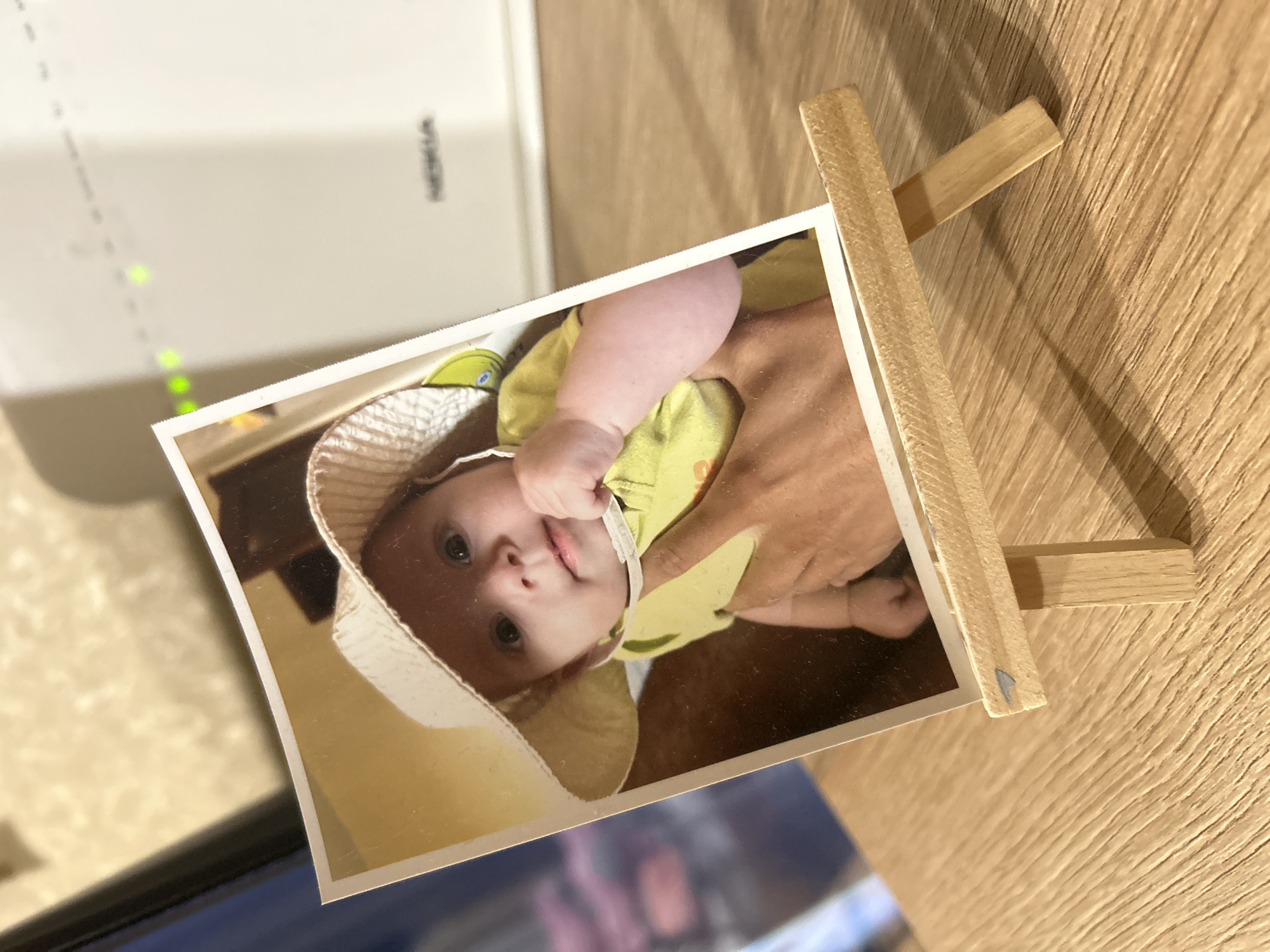 An image displays a printed photograph, oriented vertically, held upright by a small wooden easel on a light-colored wooden surface. The photograph features a baby, seemingly held by an adult whose hand is partially visible in the lower right of the photo. The baby, with light eyes and a fair complexion, is wearing a light-colored hat (possibly cream or white) and a light green or yellow top. Its small hand is near its mouth, and it gazes directly at the camera with a curious or content expression.
In the background, to the left of the easel and photo, is a white electronic device, likely a router or modem, with the brand name "NOKIA" visible. Several bright green indicator lights are illuminated on the device. The wooden surface beneath the easel shows a distinct grain pattern. Below the main photograph, a blurry reflection suggests another object or screen. The overall setting appears to be indoors, possibly a home or office, with ambient lighting.