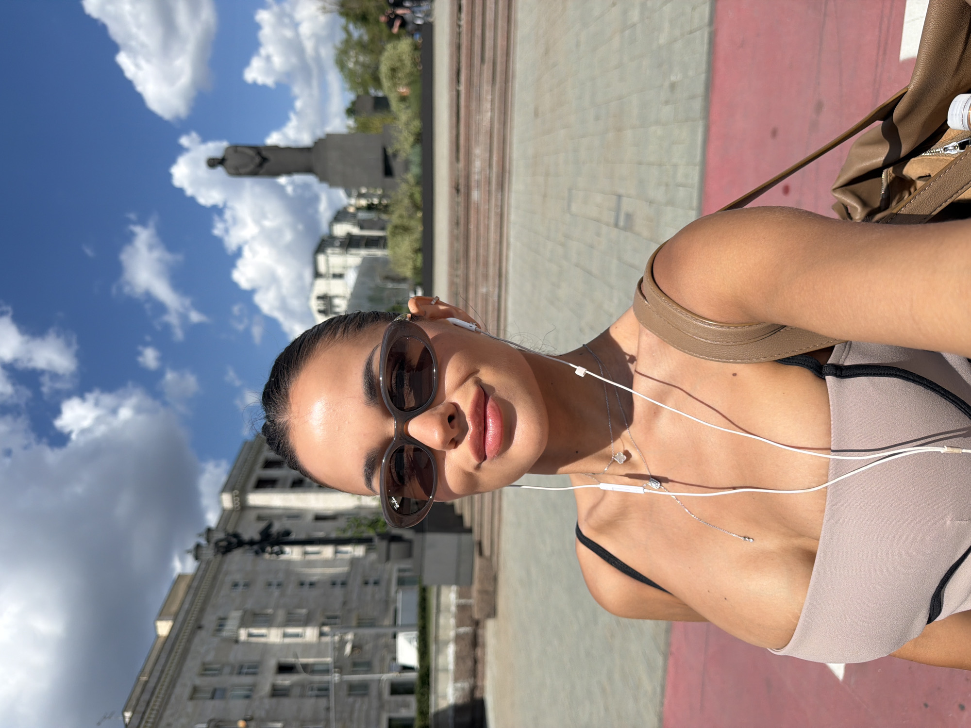 The image is an outdoor selfie featuring a young woman on a bright, partly cloudy day in what is indicated to be Moscow, Russia. The woman, positioned on the right side of the frame, looks directly at the camera with a neutral expression and a slight smile. She has dark hair styled away from her face and is wearing dark, oversized sunglasses, with the "Ray-Ban" logo visible on the right lens. White wired earphones are in her ears, their wires draped over her chest, suggesting she is listening to music. She wears a light beige top with black straps and two delicate necklaces: one with a four-leaf clover motif and another with a small, sparkly pendant. A brown, leather-like shoulder bag is slung over her right shoulder.
The background reveals an urban setting under a vivid blue sky dotted with fluffy white clouds. To the left, a large, light-colored building with multiple windows and architectural details is visible. Further back, a prominent statue of a figure on a pedestal stands against the sky. The ground consists of paved areas, some appearing as light grey tiles and others with a reddish-pink hue. The scene suggests a relaxed moment during the daytime in a city square or a wide pedestrian area.