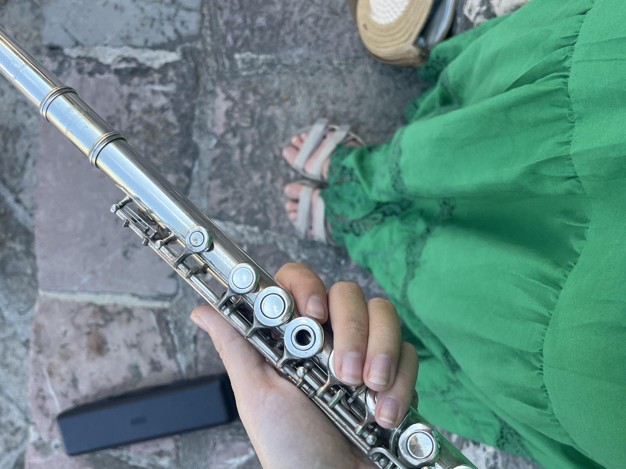 A first-person perspective shot shows a person holding a silver flute. The flute is being held in the right hand, with the fingers wrapped around the lower section of the instrument. The flute has a shiny, metallic finish and visible keys and pads. To the left of the flute, a black flute case rests on a stone surface. The person is wearing a bright green dress that drapes over their lap, and beige sandals are visible on their feet. The background consists of a stone pathway or stairs, with some weathered, reddish-brown stones. A straw hat with a light-colored brim is visible on the right side of the frame. The scene appears to be outdoors, with natural lighting. The location is identified as Budva, Montenegro.