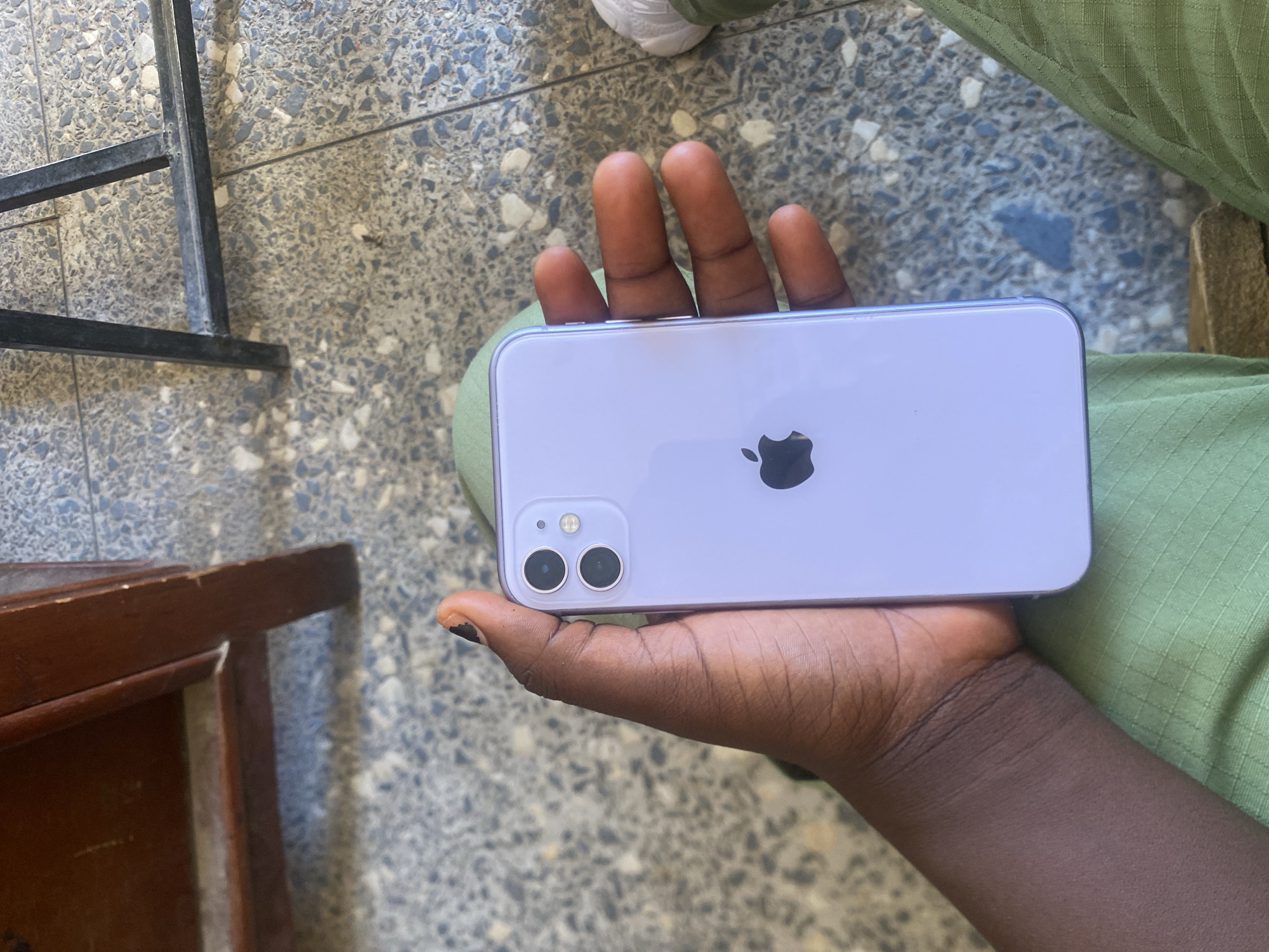 The image shows a person's hand holding a light purple iPhone 11. The phone is positioned horizontally, with the Apple logo visible in the center of the back. The camera module, featuring two lenses, is in the upper left corner. The hand appears to have dark skin and black nail polish on one finger.
The person is wearing a textured green garment, possibly pants or a skirt, which is also visible in the foreground. The background consists of a speckled terrazzo-like floor and what appears to be a section of a dark metal structure on the left side, possibly part of a table or chair. A wooden furniture piece is also partially visible on the bottom left.
The lighting suggests it is daytime, with natural light casting shadows on the floor and illuminating the objects. The scene appears to be outdoors or in a well-lit indoor space. No text is visible in the image. The overall impression is a casual, everyday scene.