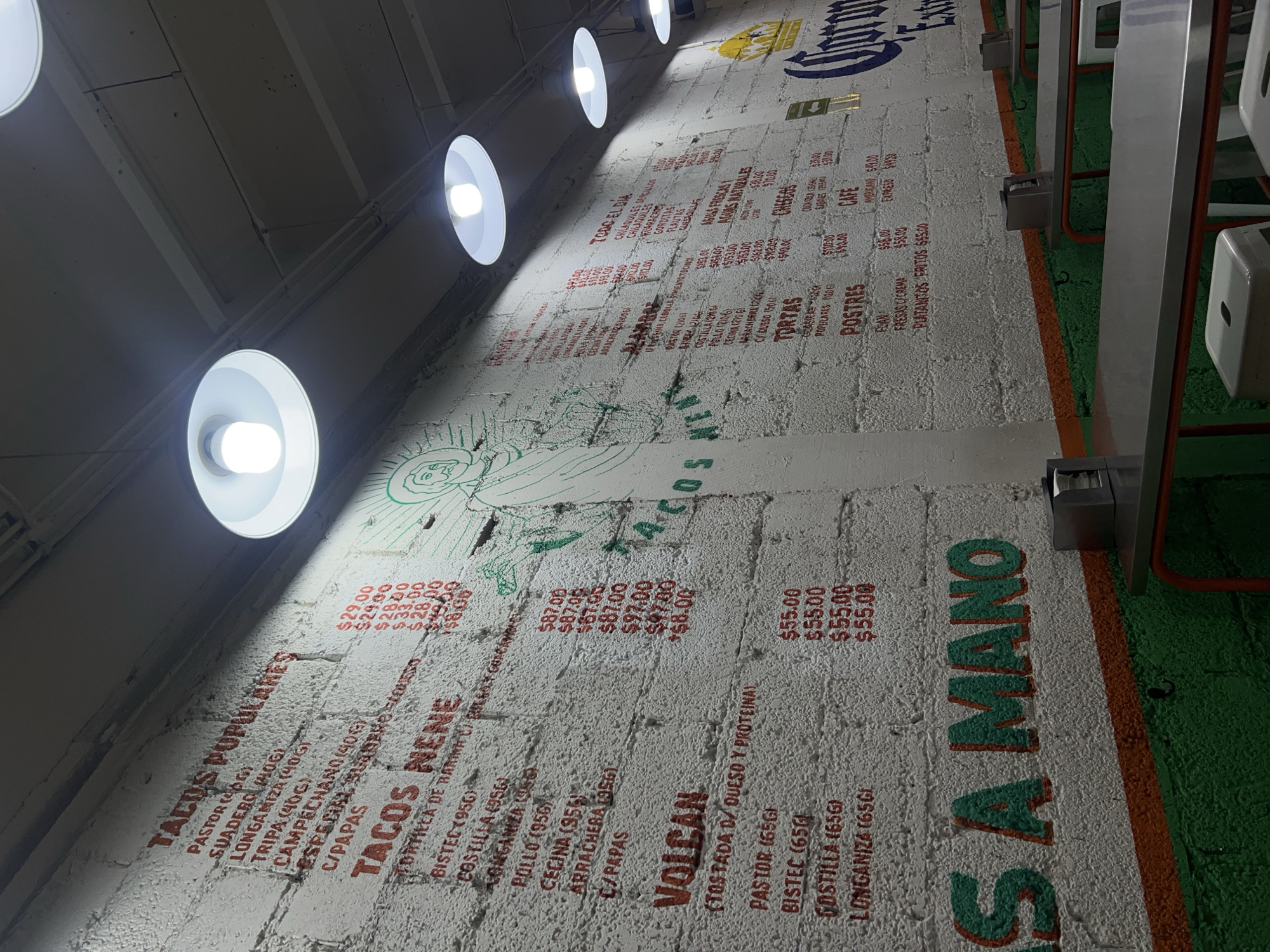 The image depicts a wall menu with food items and prices, possibly in a restaurant or food stall. The menu is written in Spanish and includes items like "Tacos Populares," "Tacos Nene," "Volcan," and "Aguas Frescas." Each item has a corresponding price in Mexican pesos. The menu is painted or printed on a textured white wall, with a Corona logo visible at the top. The perspective is from a low angle, looking up at the wall. There are bright circular lights mounted on the ceiling, and the edge of a dining area with green flooring and metal chairs is visible on the right side of the frame. The image is likely taken in Mexico City, Mexico, given the mention of Mexican pesos and Spanish menu items.