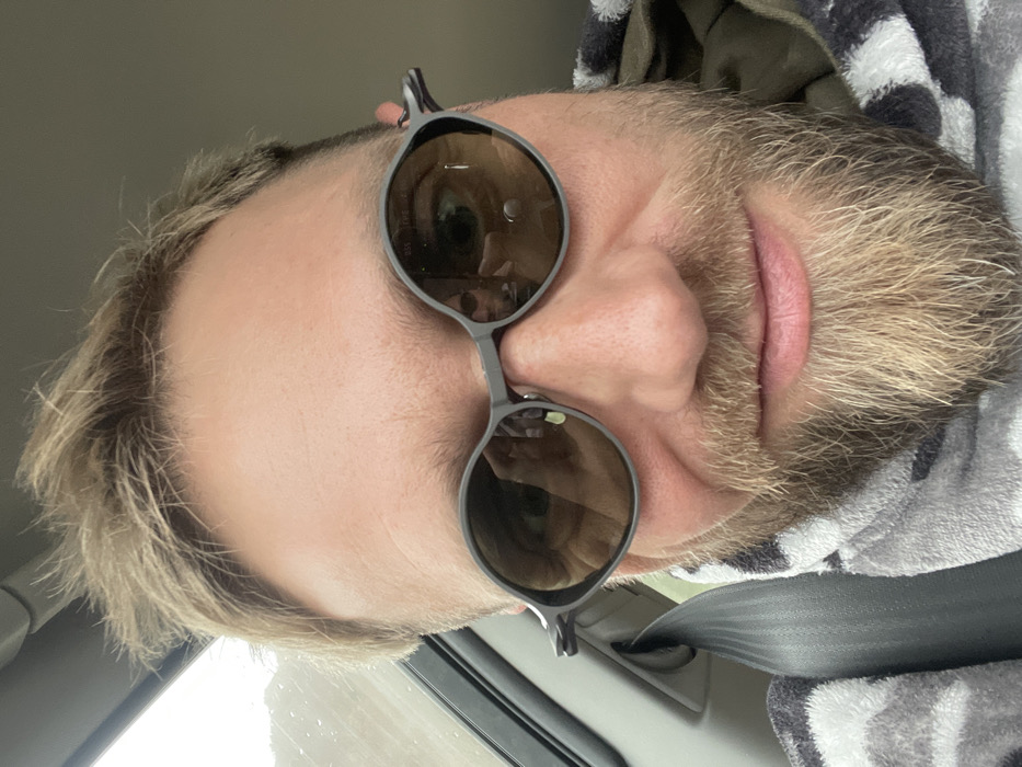 An adult male is depicted in a close-up, eye-level selfie taken from within a vehicle. He has short, light-colored hair and a full, light-blonde/ginger beard and mustache. A slight smile is present on his face. He is wearing dark-framed, oval sunglasses with amber-tinted lenses, which reflect a smartphone camera and parts of his own eyes. The male is wearing a grey and black patterned garment, possibly a hooded robe or blanket, over an olive green layer. A grey vehicle seatbelt is positioned across his left shoulder and chest. The background shows elements of a car interior, including a window frame and a blurred view through a window on the upper left, indicating an overcast sky or bright outdoor conditions. No specific landmarks or unique features are visible to confirm the location as Bukovik, Montenegro, based solely on the image content.