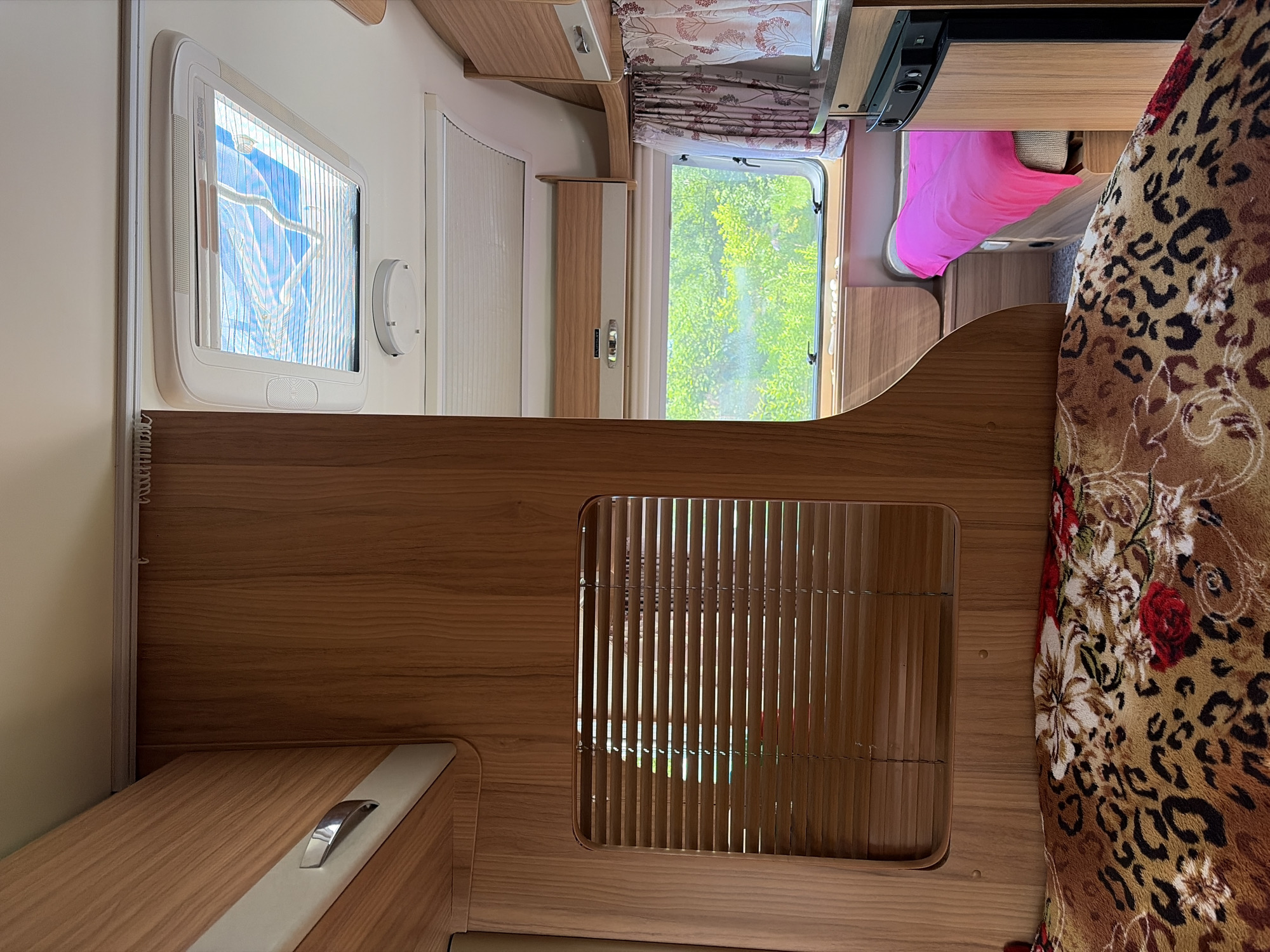 The image depicts the interior of a caravan, likely in Novozavidovskiy, Russia. The scene is dominated by wood-paneled walls and cabinets. In the foreground, a bed is covered with a blanket featuring a leopard print and floral design. To the left, a window with wooden blinds is visible. Above, a skylight with blinds is open, revealing a blue sky and some fabric. A larger window in the mid-ground looks out onto a lush green landscape with trees. A bed with a bright pink blanket is visible in the distance. A small black refrigerator is situated to the right. The overall impression is that of a compact living space within a recreational vehicle.