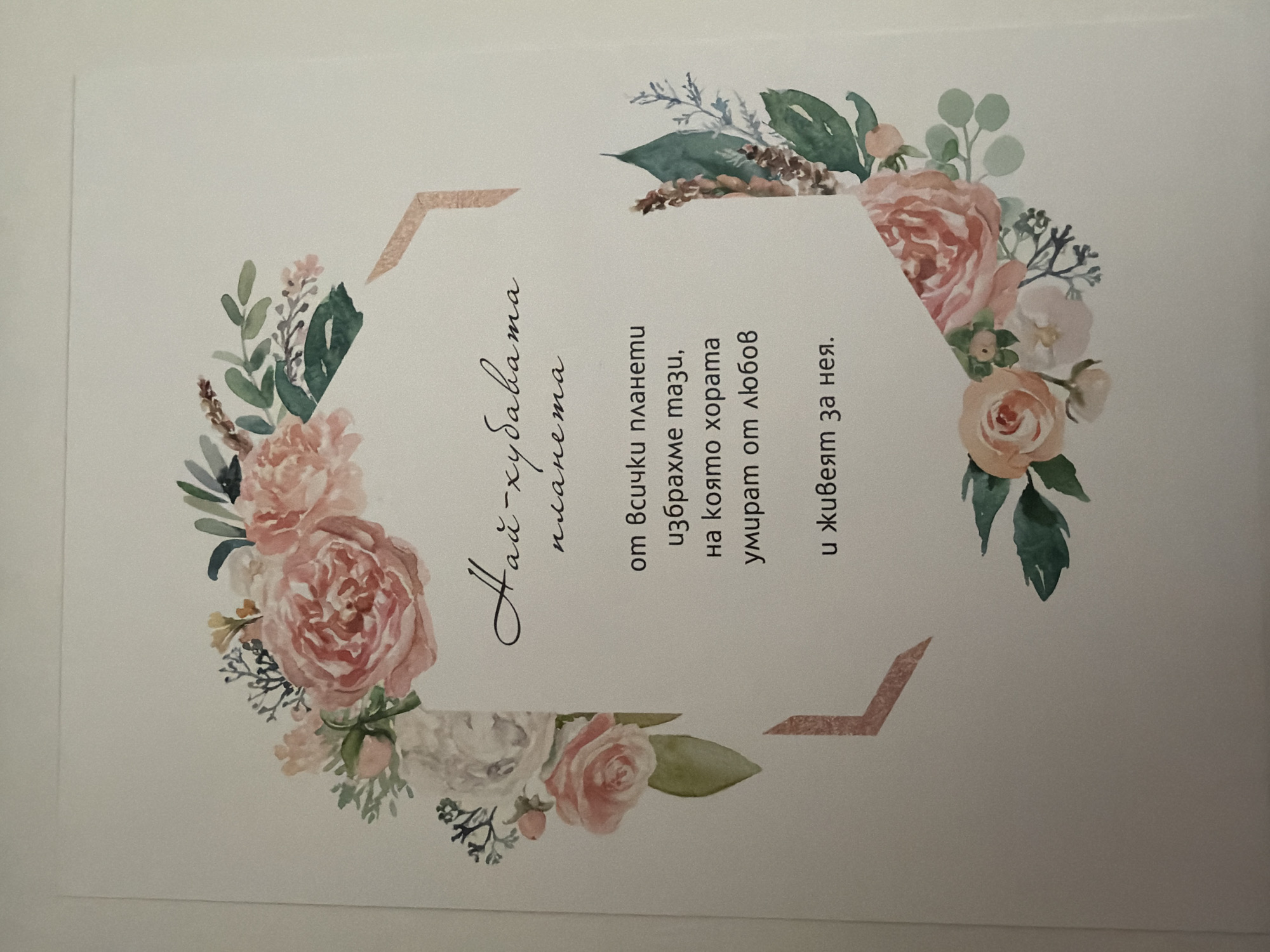 The image displays a card with a floral watercolor design. The card has a light background and is decorated with a wreath of pink and white roses and green foliage. In the center of the card, there is text written in Bulgarian. The text reads: "Най-хубавата песен" followed by "от всички планети избрахме тази, на която хората умират от любов и живеят за нея." This translates to "The most beautiful song" followed by "from all the planets we chose this one, where people die for love and live for it." There are no people, animals, or other objects visible in the image. The card is placed on a plain white surface, which could be a table or a wall. The location of this image in the city of Pazardzhik, Bulgaria, is not provided in the image itself.