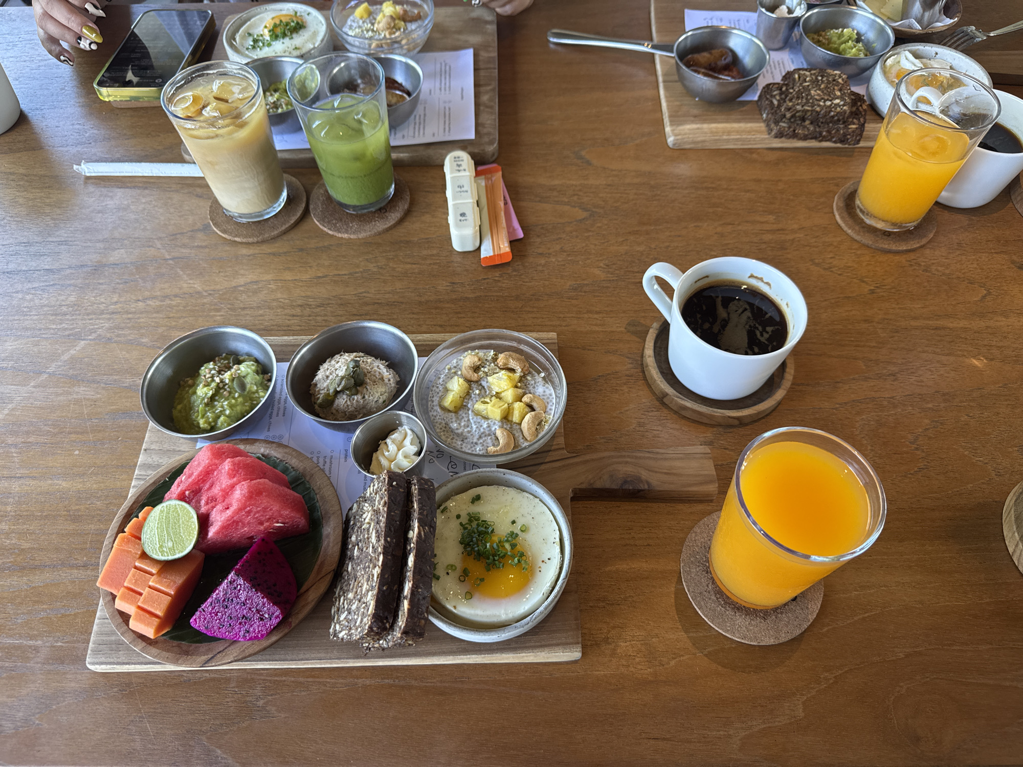 This photo presents a wooden table laden with various food and beverages, seemingly in a restaurant in North Kuta, Indonesia. Two sets of meals can be distinguished, likely for two people. Each set includes several small metal bowls containing diverse dishes, such as guacamole, a spread with seeds, chia seed pudding with fruit, and an egg dish. Alongside these are slices of dark bread, cut fruits (watermelon, papaya, dragon fruit, and lime), and a small container of cream. The beverages include coffee and orange juice, each placed on a round coaster. One set also has an iced coffee and a green juice. Near the iced coffee and juice is a pill organizer and a couple of candy wrappers. A cell phone is also seen on the edge of the table near one of the place settings.