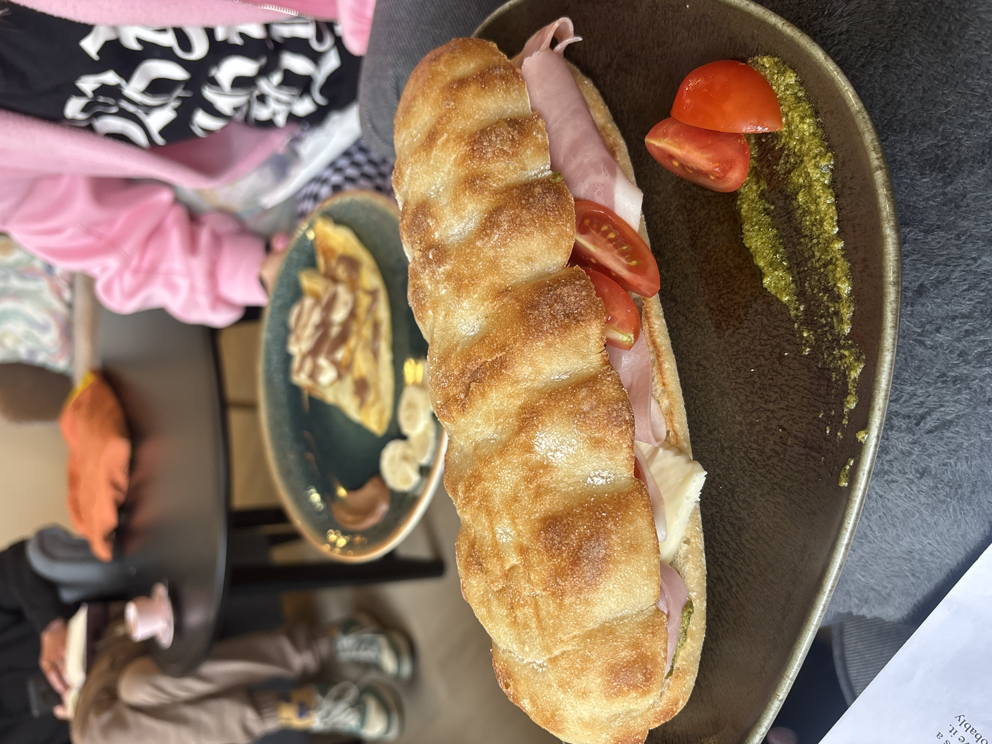 The image depicts a sandwich on a brown plate, accompanied by tomato slices and pesto sauce. The sandwich appears to be made with a baguette-style bread, ham, and cheese. In the background, there is another plate with a dessert, possibly a crepe or pancake with a brown sauce and sliced bananas. In the blurred periphery of the shot, there is some clothing and a table setting, suggesting an indoor dining environment, possibly in a café or restaurant. There is no information in the picture to verify the location in the city of Varna, Bulgaria.