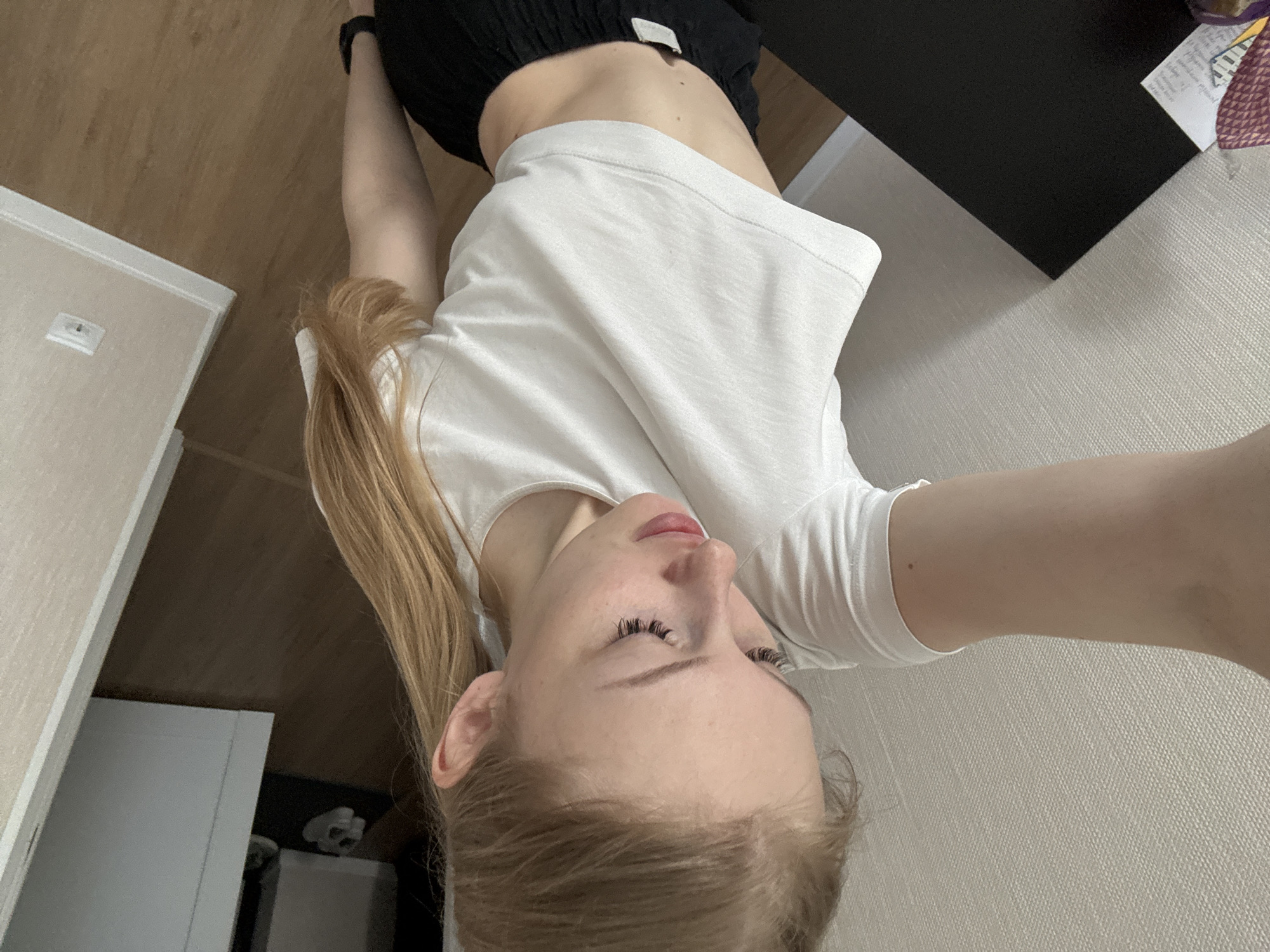 The image features a young woman with blonde hair, wearing a white t-shirt and black shorts. She is positioned in a way that suggests a selfie is being taken, with her face tilted upwards and her arm extended. The background shows a room with wooden flooring and a wall with a textured finish. There is also a glimpse of what appears to be furniture and a doorway. The lighting suggests it is daytime, but no specific time can be determined. The overall mood is casual and relaxed. No visible text is present in the image. The location context is Perm, Russia.