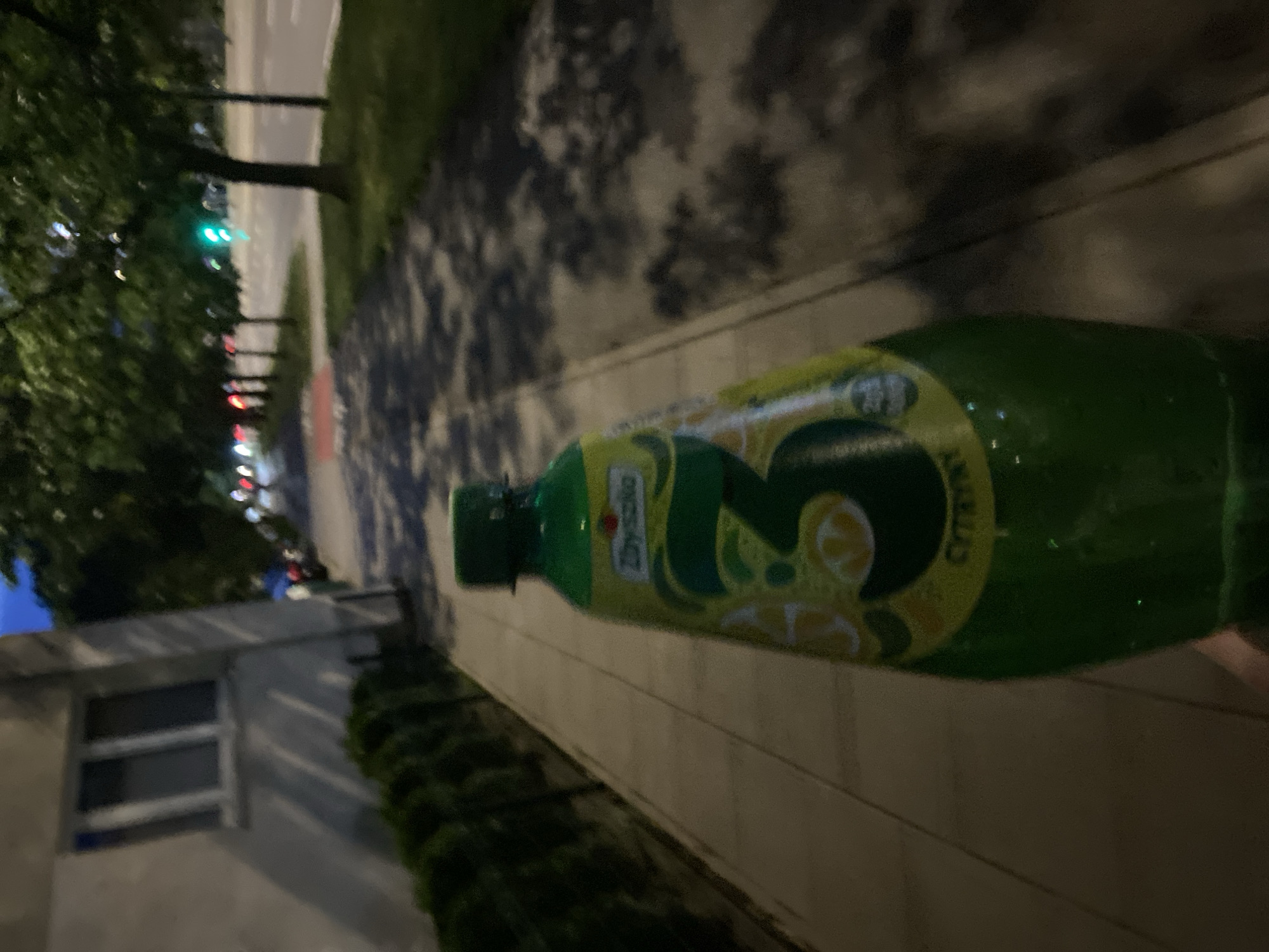 The image shows a green plastic bottle of "3 Cytryny" (3 Lemons) drink, held by an unseen person, in what appears to be an urban environment. The bottle has a label with a large "3" and images of lemons. In the background, there is a sidewalk lined with green bushes, a building with windows on the left, and a street with a traffic light showing green on the right. Trees are visible along the street. The lighting suggests it is dusk or evening. The overall scene is consistent with a street in Warsaw, Poland, given the presence of the Polish brand and typical urban elements.