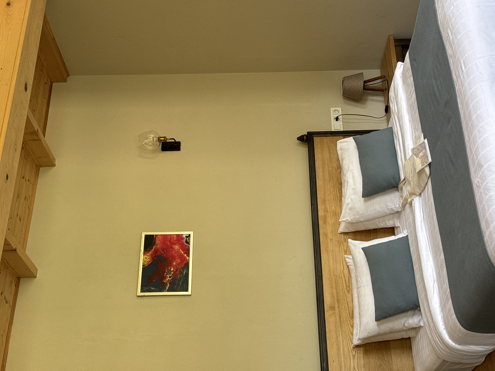 The image depicts a hotel room in Tibeta, Georgia. The scene features a bed with white linens and a dark grey runner, adorned with two white pillows and two dark grey accent pillows. On the bed, a beige tote bag with some papers and a small framed photo is visible. The headboard is made of wood and black metal. To the right of the bed, a small wooden nightstand holds a lamp with a conical shade. The wall behind the bed is painted a light olive green and features a framed abstract painting with red and gold tones, and a wall-mounted light fixture with a clear bulb. Above the wall, exposed wooden beams are part of the ceiling. A power outlet is visible on the wall to the right of the bed.