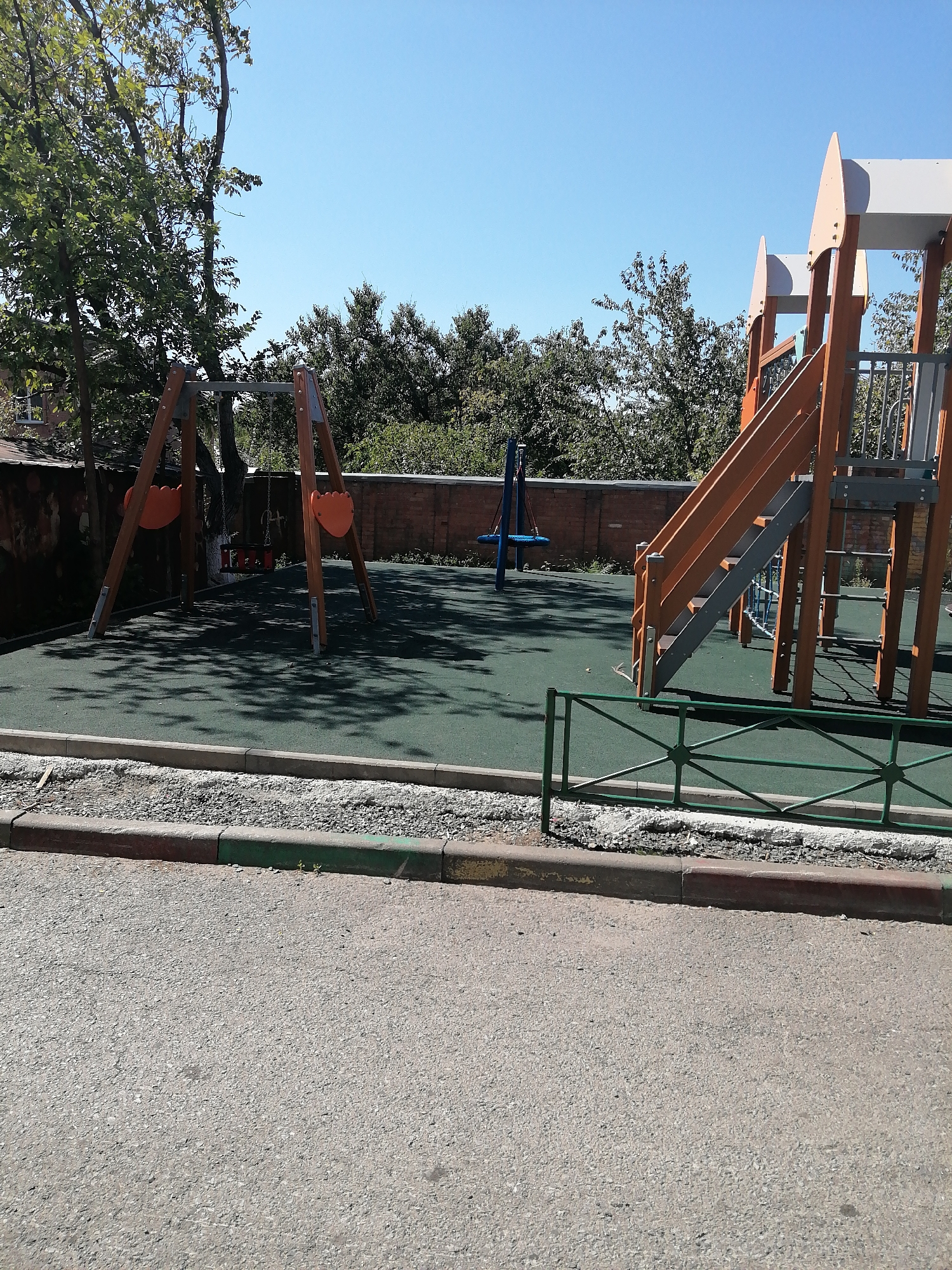 The image shows a children's playground located in a city, possibly Rostov-on-Don, Russia. The playground features a wooden swing set with two orange seats, a blue tire swing, and a large wooden structure with an orange slide and stairs. The ground surface is a green rubber mat, and the playground is surrounded by a low green metal fence. In the background, there are trees, a brick wall, and a building. The sky is clear and blue. The image was taken on a sunny day, as indicated by the shadows cast on the ground.