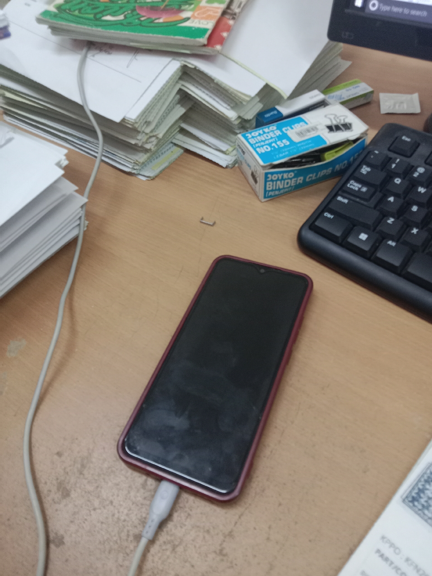 The image depicts a close-up, overhead view of a wooden desk surface. In the foreground, a dark-screened smartphone with a maroon case is plugged in and charging via a white USB cable. To the right of the phone, a portion of a black computer keyboard is visible. In the middle ground, a small blue and white cardboard box labeled "JOYKO BINDER CLIPS NO. 155" is situated, along with a stack of white envelopes and papers to its left. A light gray cable, likely a charging cable or wire, runs across the desk from the left side towards the smartphone. In the upper left corner, a stack of books with green covers is partially visible. The overall environment appears to be an office or study space. There are no people visible in the image. There is no information to suggest this scene is located in Semarang, Indonesia.