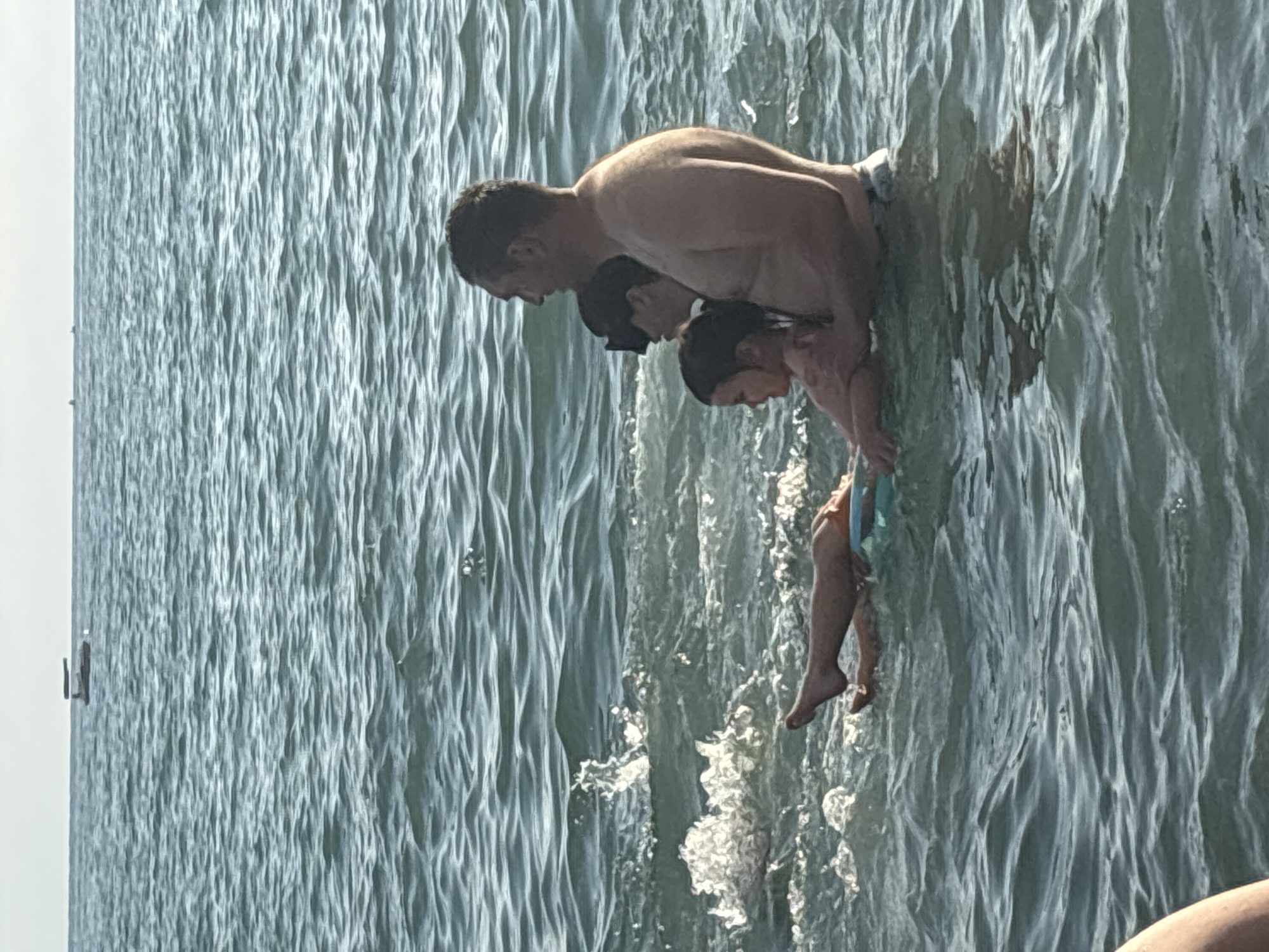 The image shows three people in the water, with a man in the foreground. He is holding two children. The water is choppy and appears to be the sea. The lighting suggests it is daytime, possibly in the late afternoon due to the shadows. The people are dressed in swimwear. The man's torso and shoulders are visible, and he is looking down at the children. The children are positioned in front of him, with one appearing to be held by the man. The water is reflecting sunlight, creating a shimmering effect. The overall scene depicts a family enjoying time together in the water. The context provided indicates the location is Sochi, Russia. No text is visible in the image.
