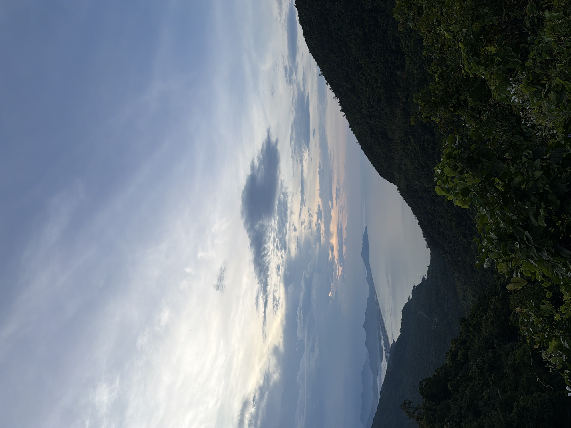 The image captures a view of the landscape, likely in a mountainous area near Huế, Vietnam, given the vegetation and water features. The sky occupies a significant portion of the image, filled with clouds ranging in color from white to dark gray. Some clouds are illuminated by the sun. A coastline or lake stretches between the mountains. The mountain is covered in vegetation. The picture has an atypical angle, and it appears to have been rotated counter-clockwise.