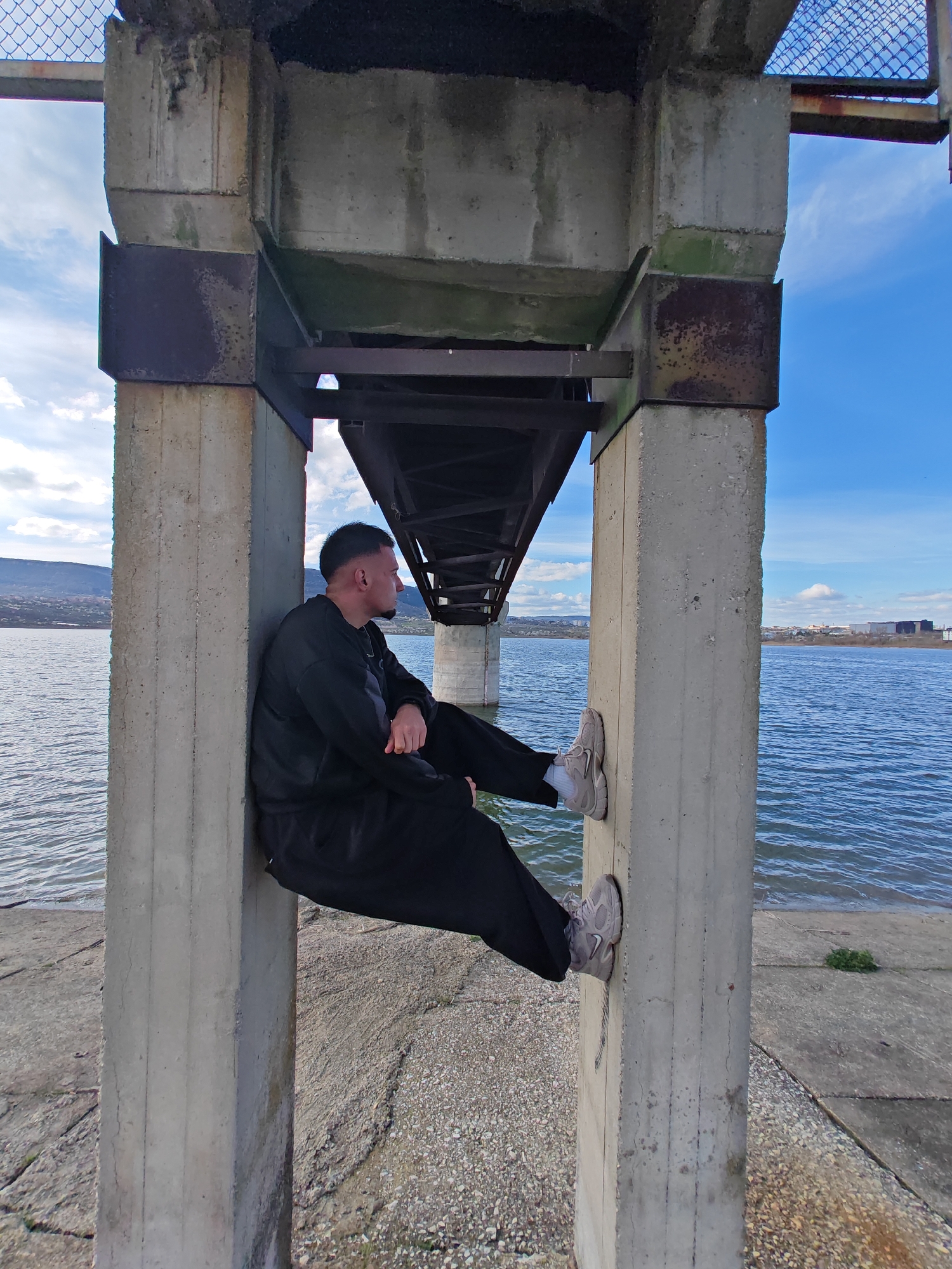A man is sitting between two concrete pillars under a bridge, appearing to rest. He is wearing a black sweatshirt and sweatpants and grey shoes. He is resting his feet against one pillar and leaning his back against the other. The bridge structure is overhead, with a metal frame visible above him. In the background is a body of water, with the opposite bank visible, and a blue sky with scattered clouds. The scene is likely located in or near Shumen, Bulgaria, judging by the terrain.