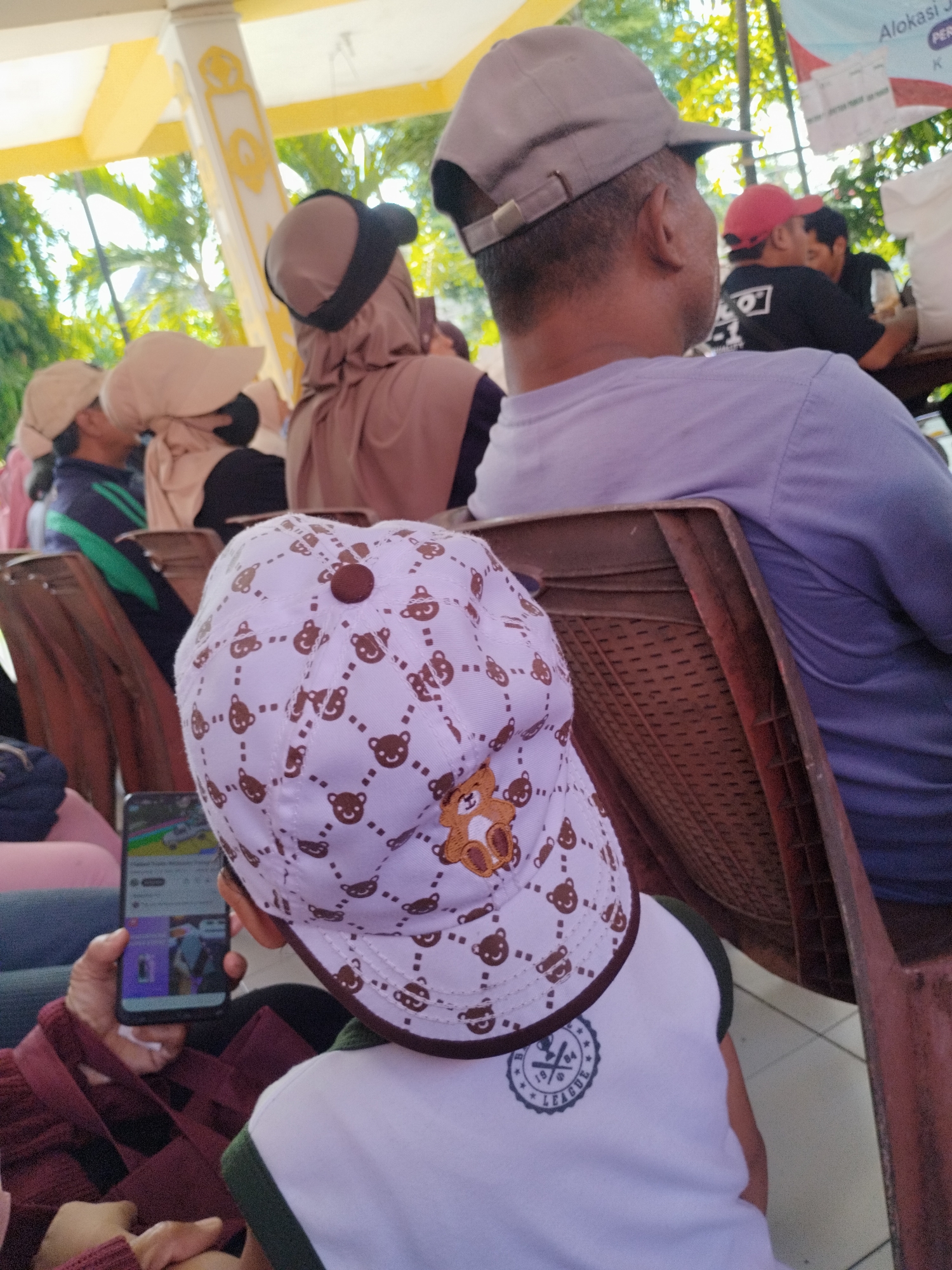 A young child wearing a white baseball cap with a bear pattern is sitting in a brown plastic chair. The cap has a brown button on top and a brown brim. The child is looking at a smartphone held by an unseen person in front of them, displaying a YouTube video. The child is also wearing a white shirt with a dark green trim and a circular emblem on the chest. The background shows other people seated, some wearing head coverings. The environment appears to be outdoors with trees visible, and a banner with text is partially visible in the upper right corner, suggesting a public gathering or event. The scene is set in Surabaya, Indonesia, as indicated by the presence of a banner that appears to be an advertisement or announcement for an event occurring within the city. The overall atmosphere suggests a casual, possibly crowded, event where people are waiting or observing.